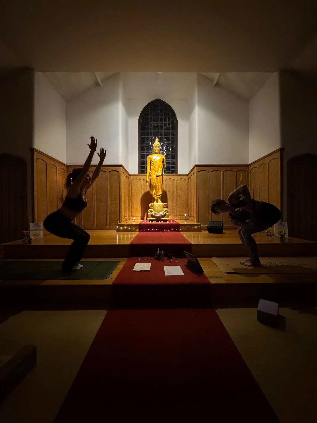 Two people practicing yoga poses in a dimly lit room with a Buddha statue on a platform at the back, a red carpet leading to the statue, and wooden paneling on the walls.