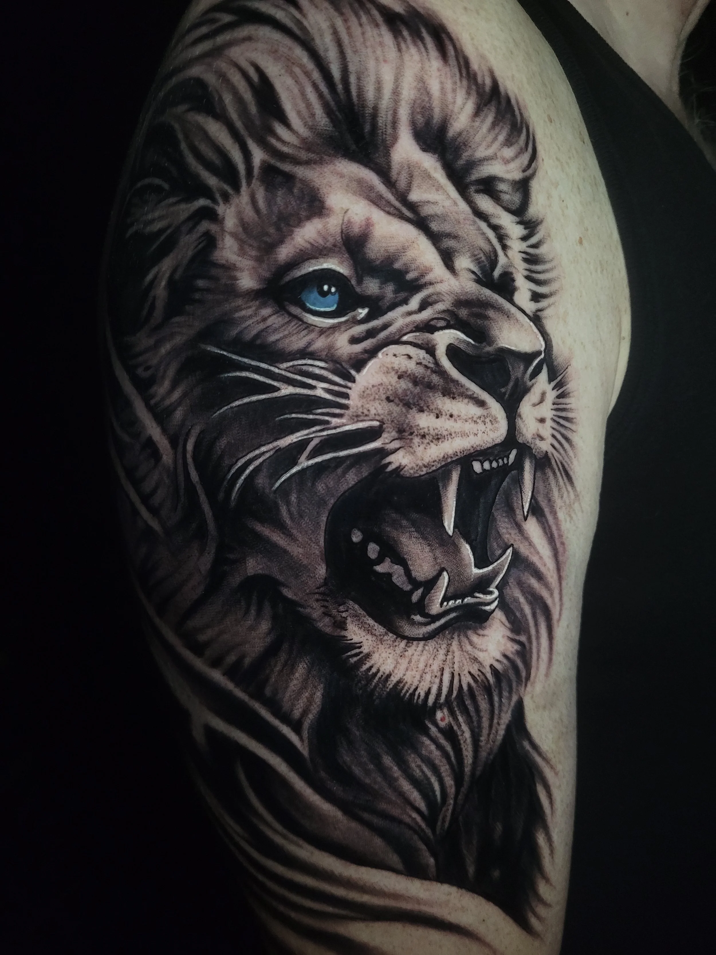 Black and gray tattoo of a roaring lion with a piercing blue eye, on a person's arm.