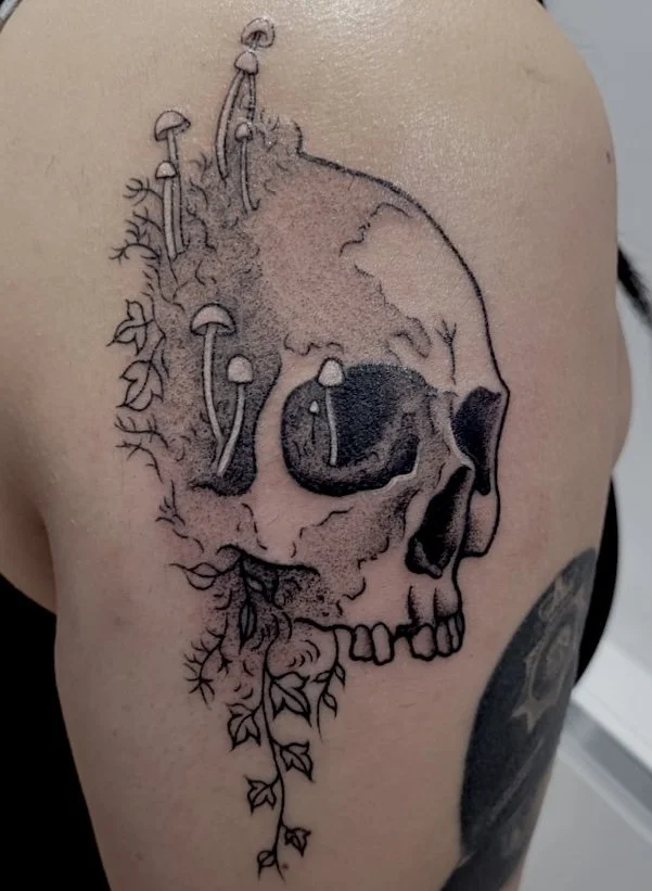 Black ink tattoo of a human skull with mushrooms and ivy growing around and from it on a person's shoulder.