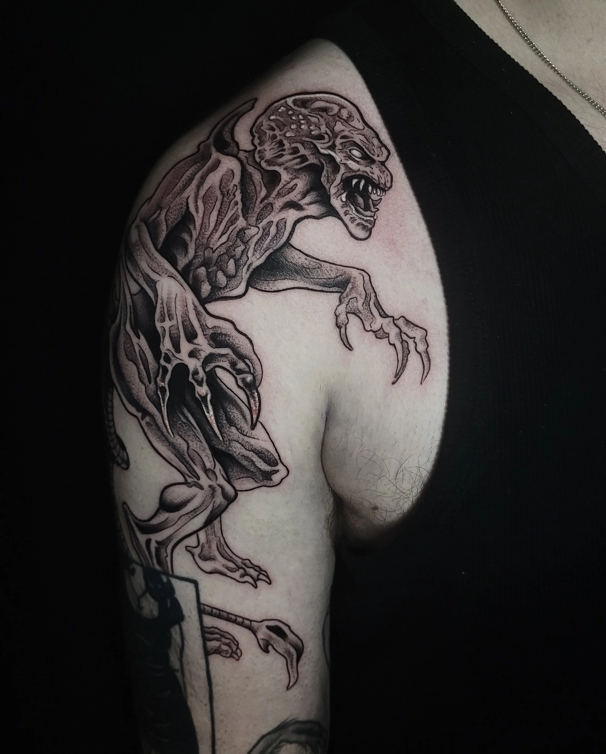 Black and gray tattoo of a monstrous creature with sharp claws, jagged teeth, and a fierce expression on a person's upper arm and shoulder.
