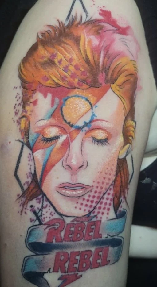 Tattoo of a woman with closed eyes, vibrant makeup, and a colorful style resembling David Bowie, with the words 'Rebel Rebel' beneath.