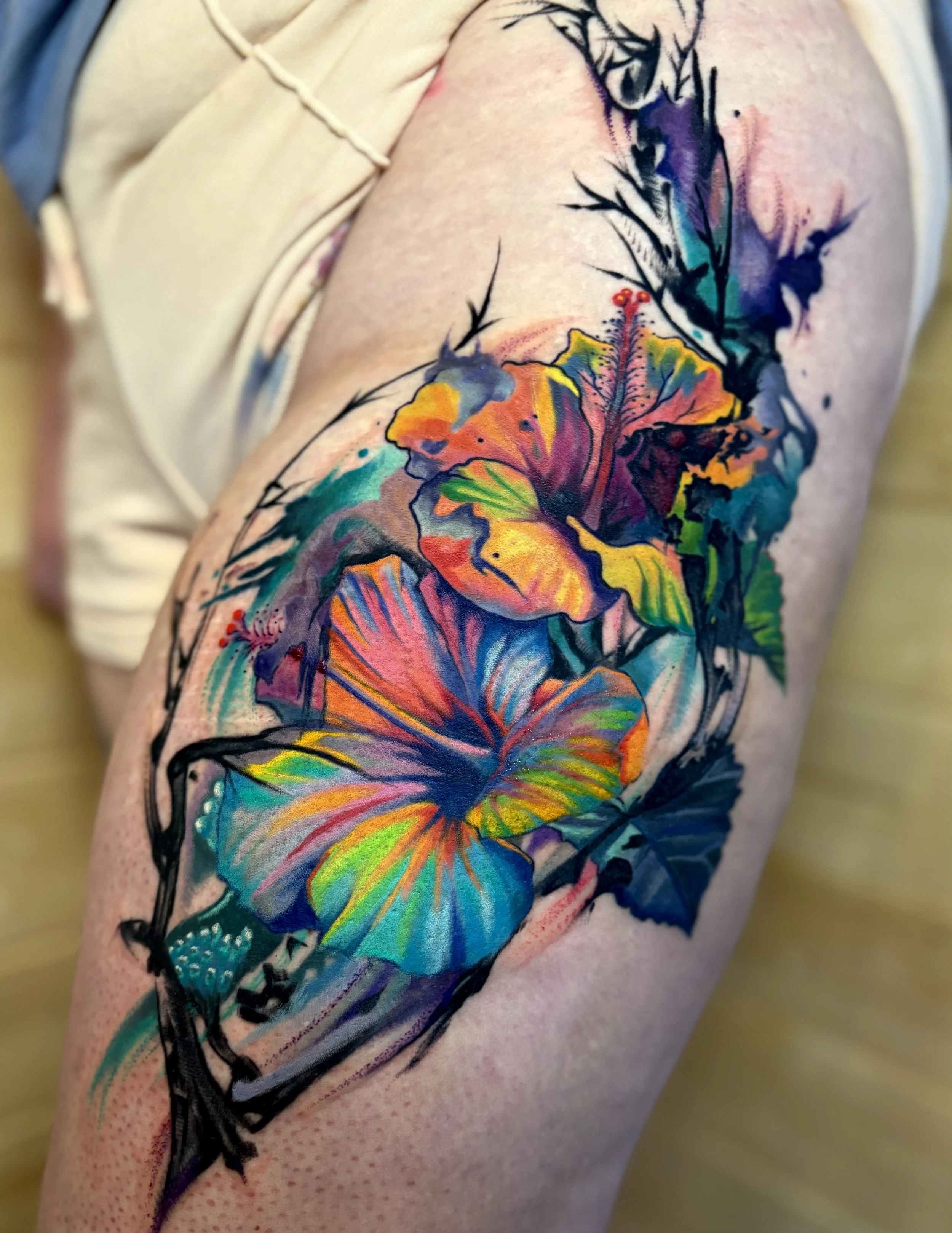 Colorful watercolor-style floral tattoo featuring large hibiscus flowers and leaves on upper arm.