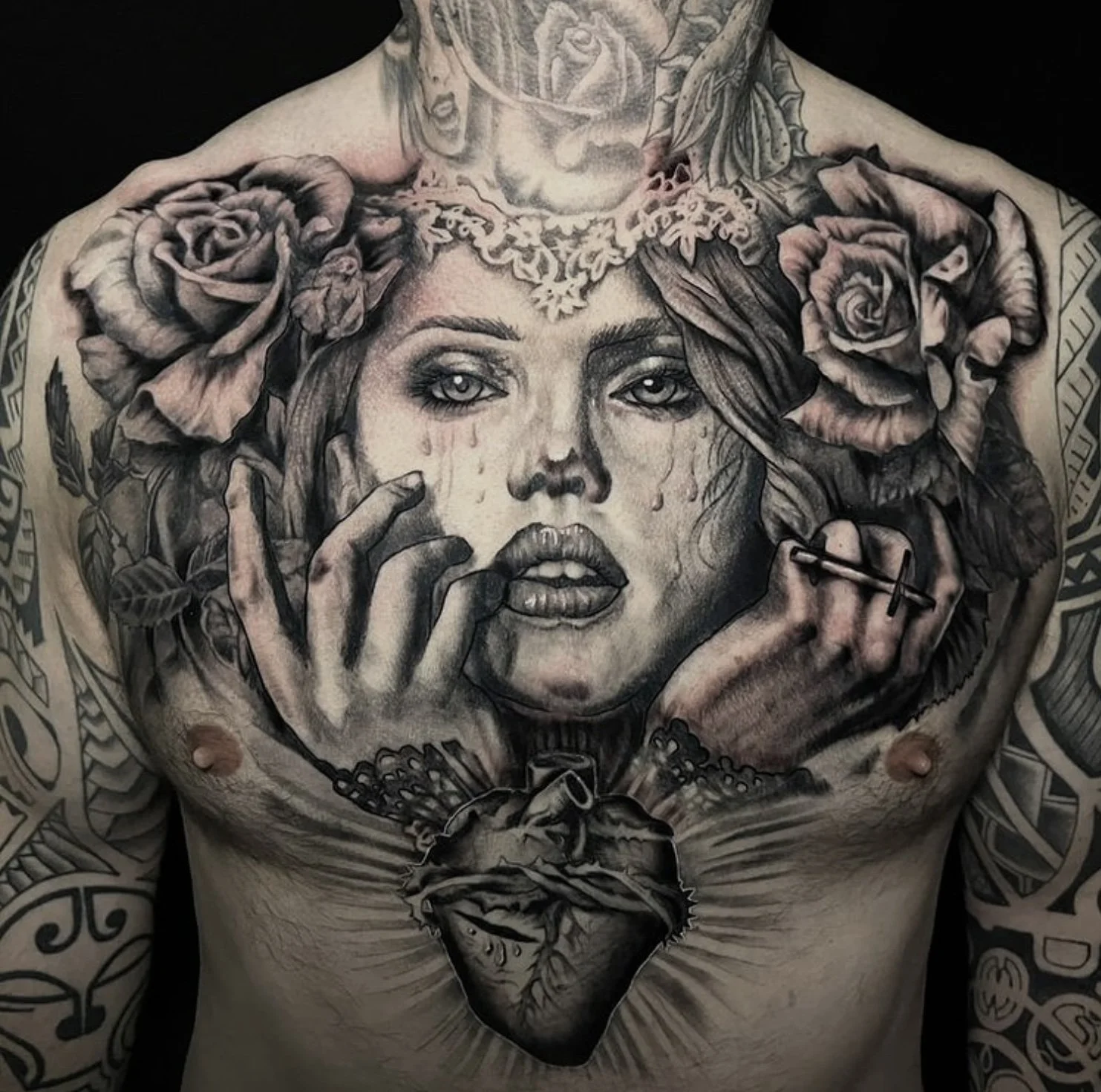 Black and gray tattoo of a woman's face with tears, surrounded by roses, lace, and ornamental designs, with a heart on the chest.