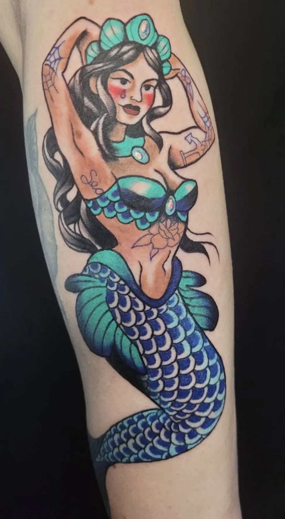 Tattoo of a mermaid with long black hair, teal seashell top, and blue-green scales on her tail, with a tattooed flower on her abdomen, and a tear drop on her face.