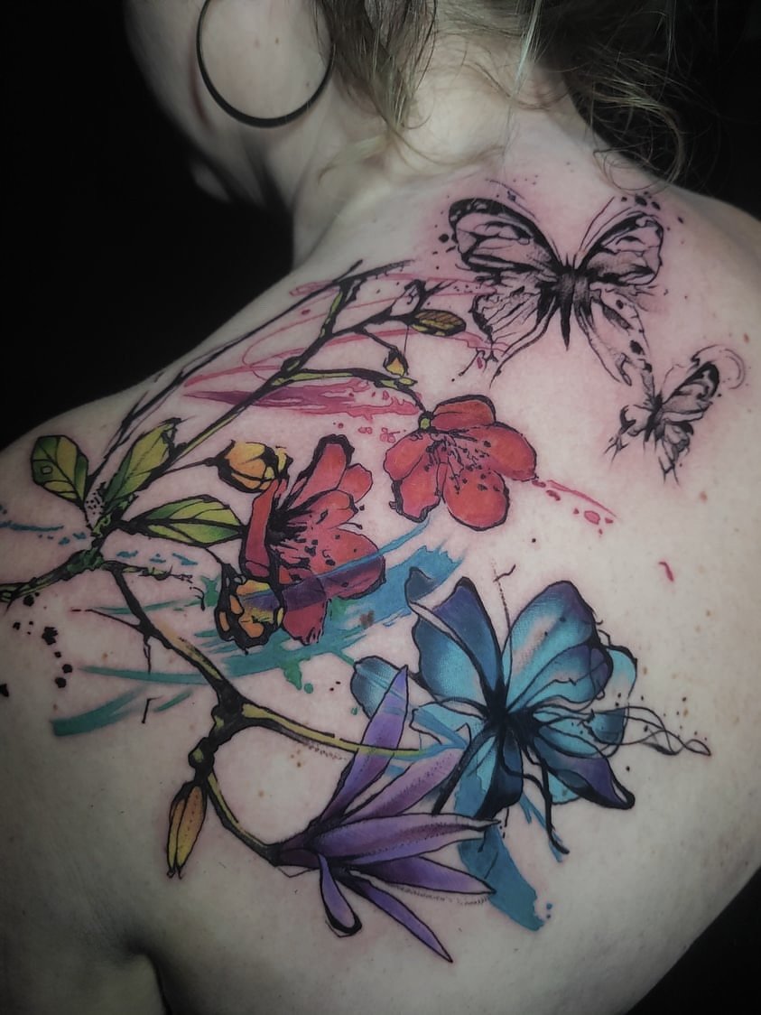 Colorful tattoo of butterflies and flowers on a person's shoulder and chest, with watercolor-style splashes.