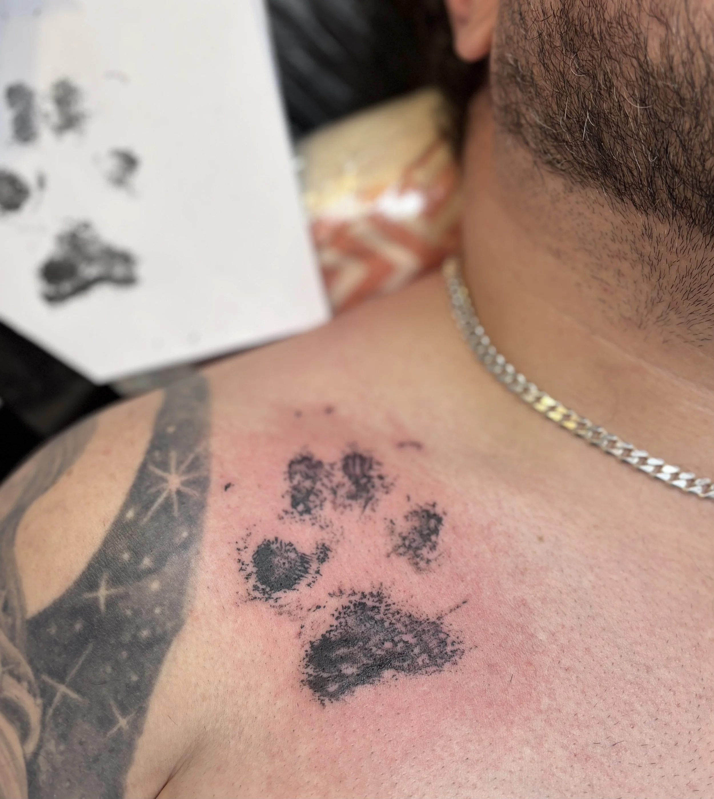 Tattoo of animal paw prints on person's shoulder and neck, with some smudged ink and redness around the tattoo; person has a silver chain necklace and short facial hair.