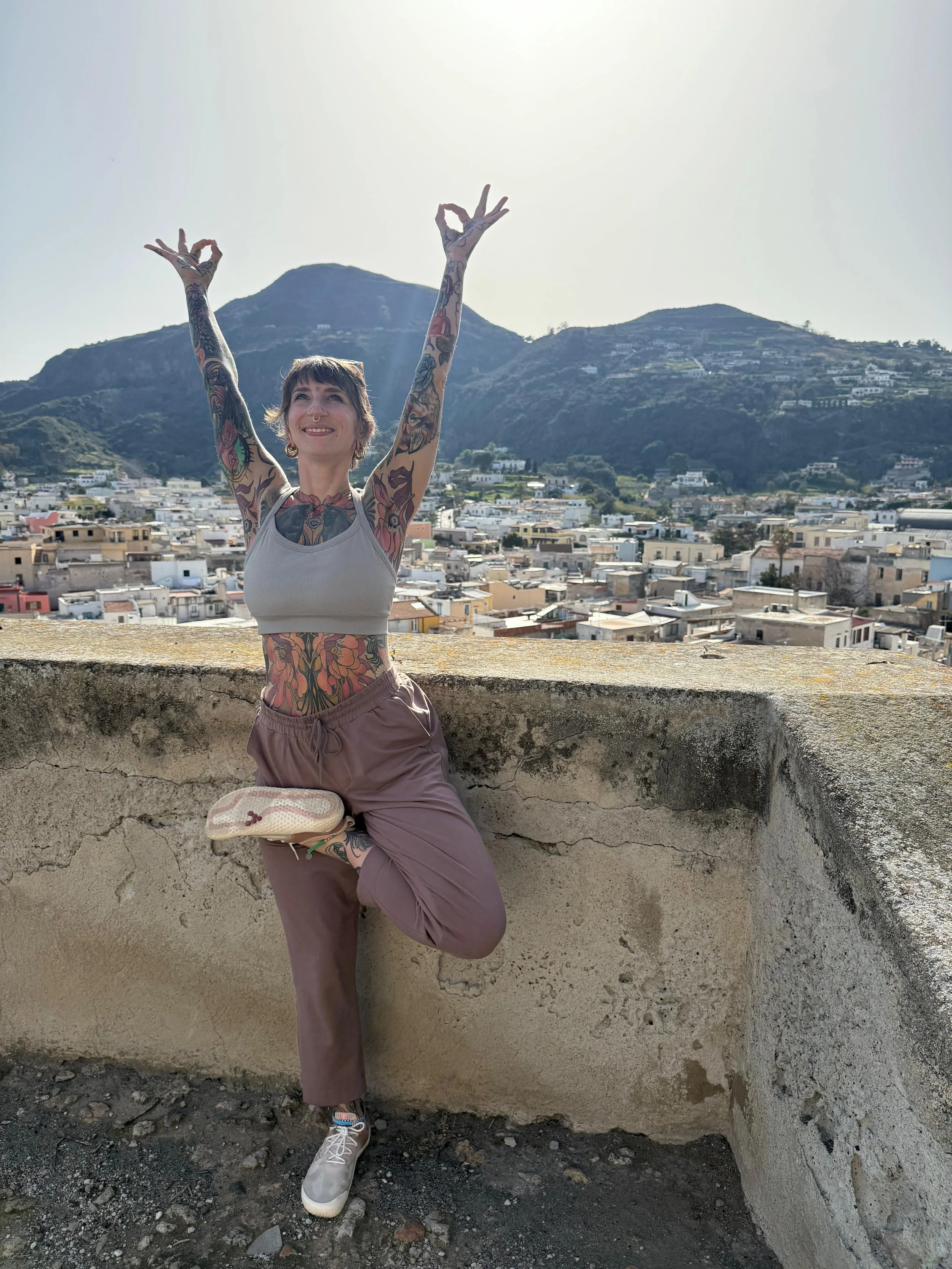 A woman with tattoos on her arms and torso is standing on one leg in a yoga pose, smiling with arms raised and making an 'OK' sign with her fingers. She is outdoors on a rooftop with a city and mountains in the background, wearing a light gray tank top, taupe pants, and sneakers.
