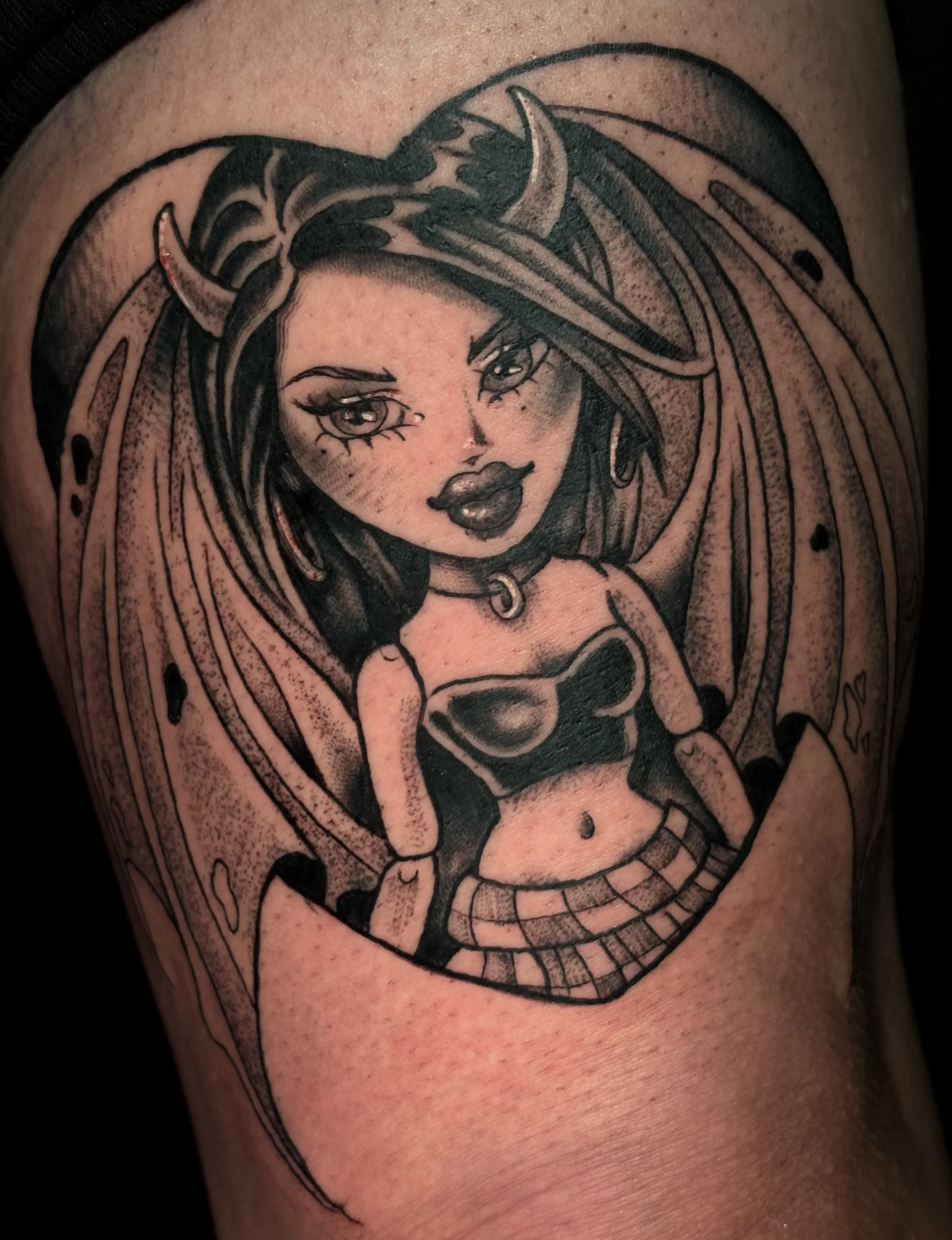 Black and gray tattoo of a female character with horns, long hair, and large eyes. She has a confident expression and wears a choker, a crop top, and a plaid skirt, with bat wings in the background.