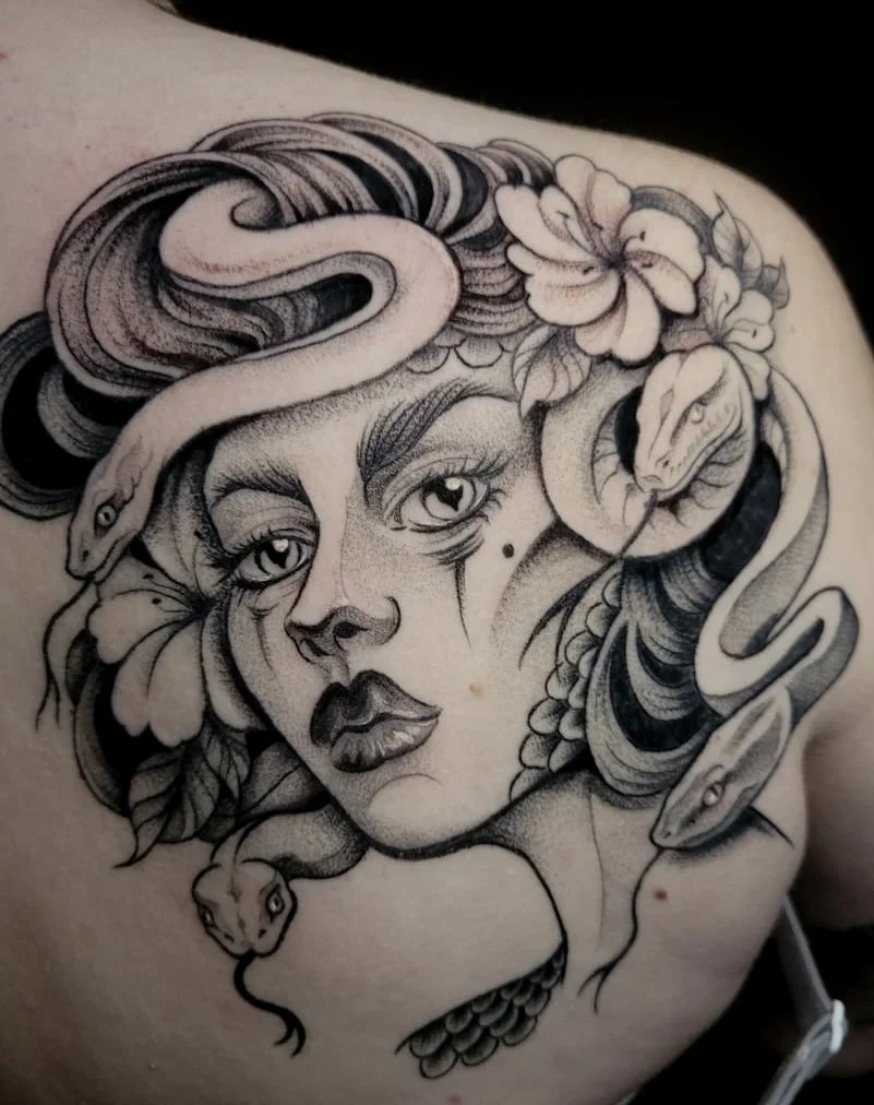 A black and gray tattoo of a woman's face with large eyes, full lips, and flowing hair intertwined with snakes and flowers.