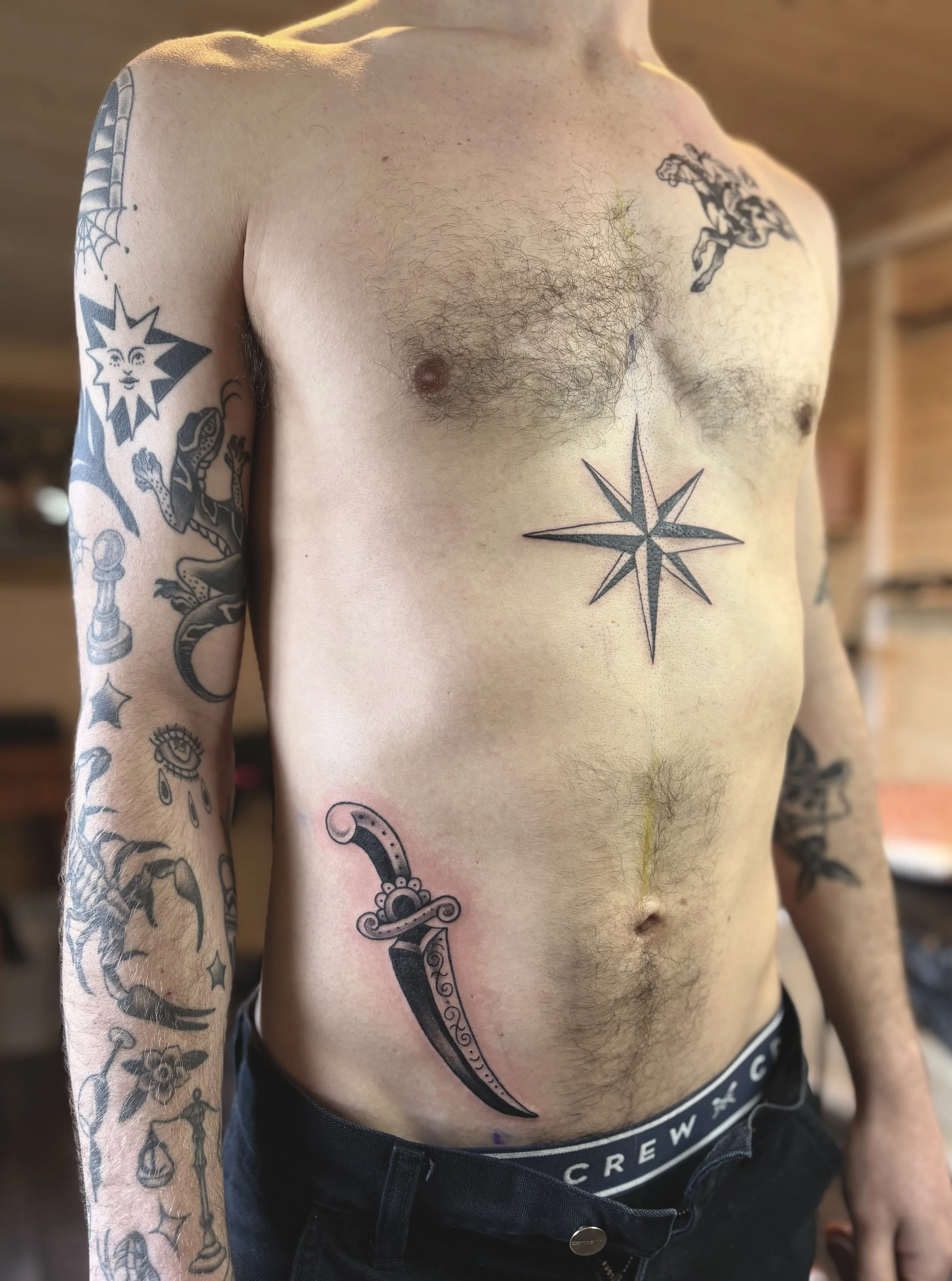 A shirtless man with tattoos on his chest and arms, including a compass on his chest, a sword on his side, and various other designs on his sleeves. The background appears to be a wooden interior.