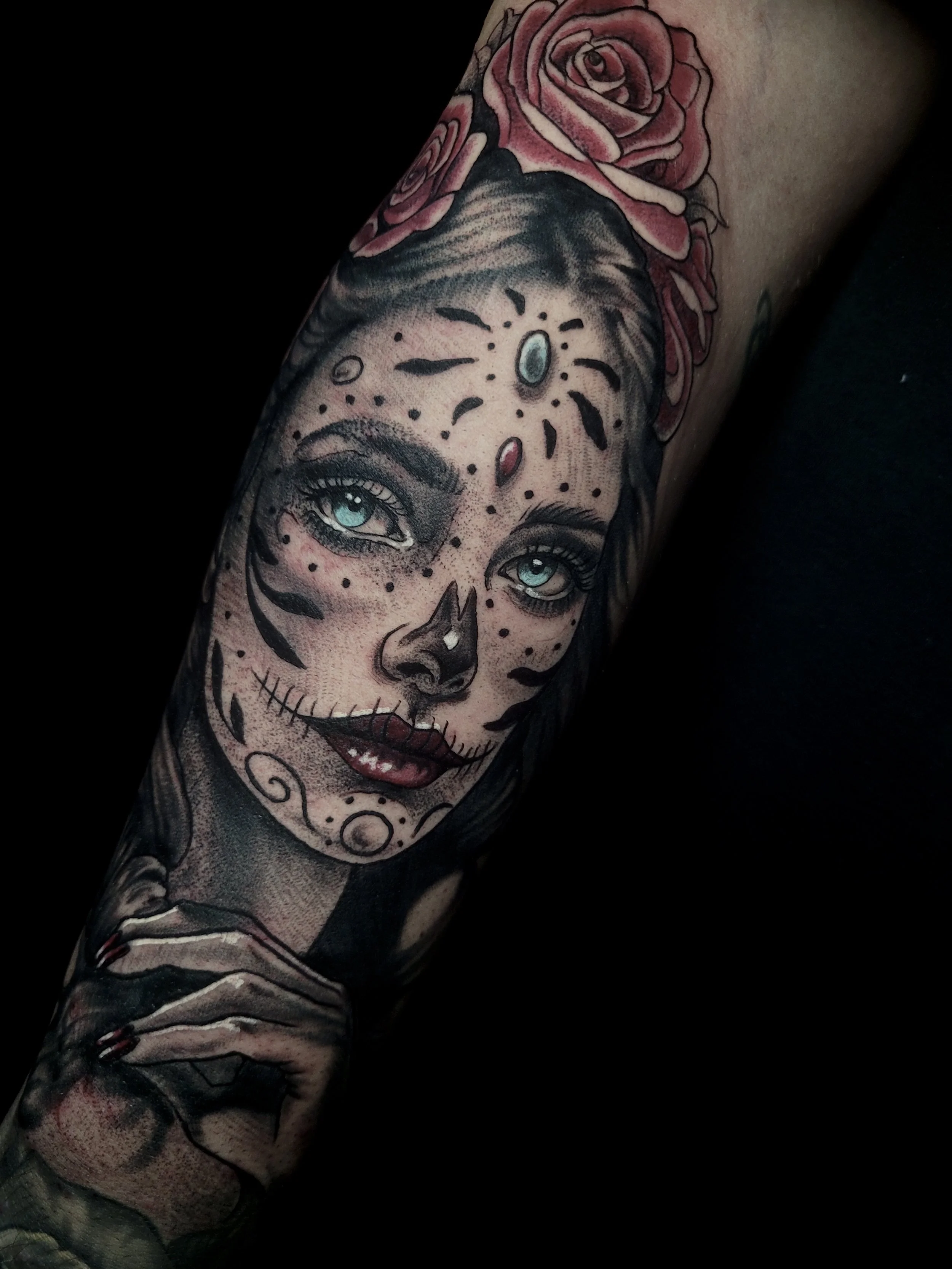 Tattoo of a woman with day of the dead makeup, blue eyes, and pink roses on her head.