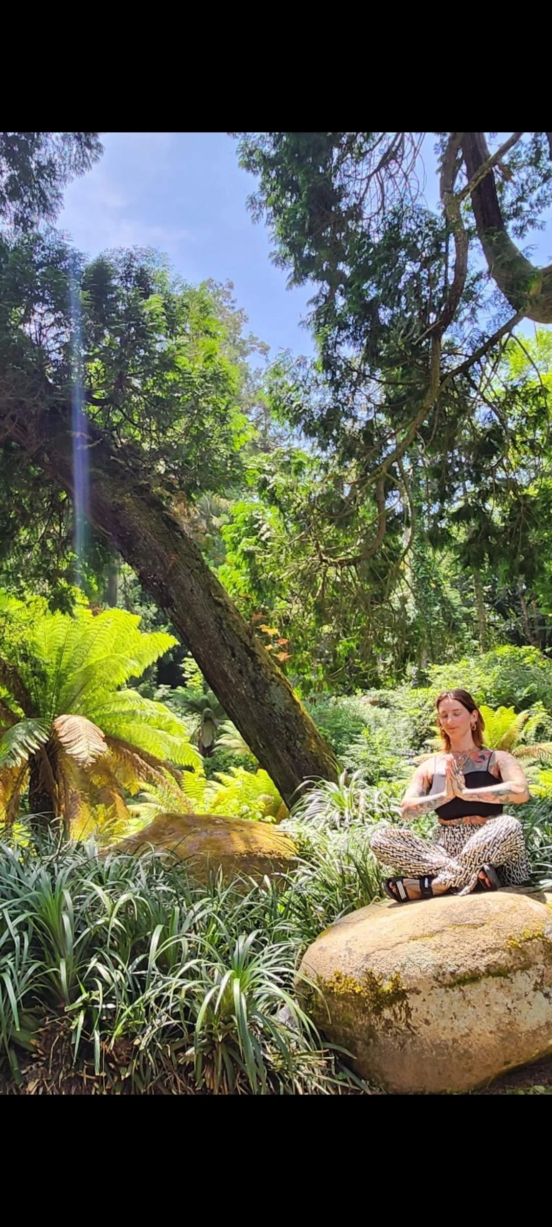 A woman practicing yoga or meditating outdoors in a lush, green forest with large rocks, dense foliage, and a clear blue sky.