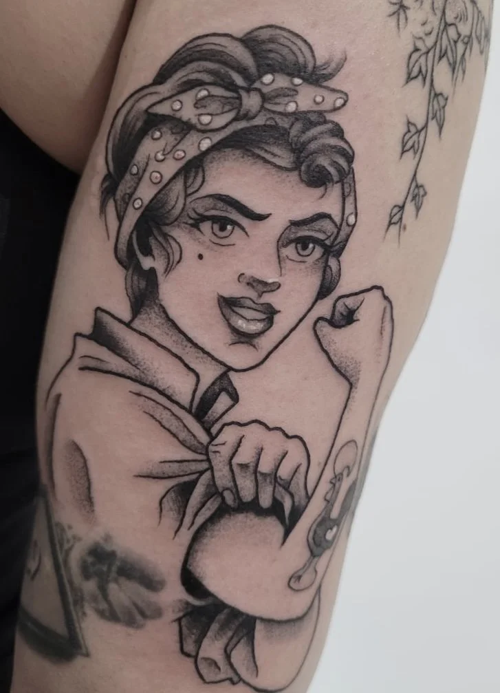 Black and gray tattoo of a woman in Rosie the Riveter pose, with a bandana on her head, flexing her arm.