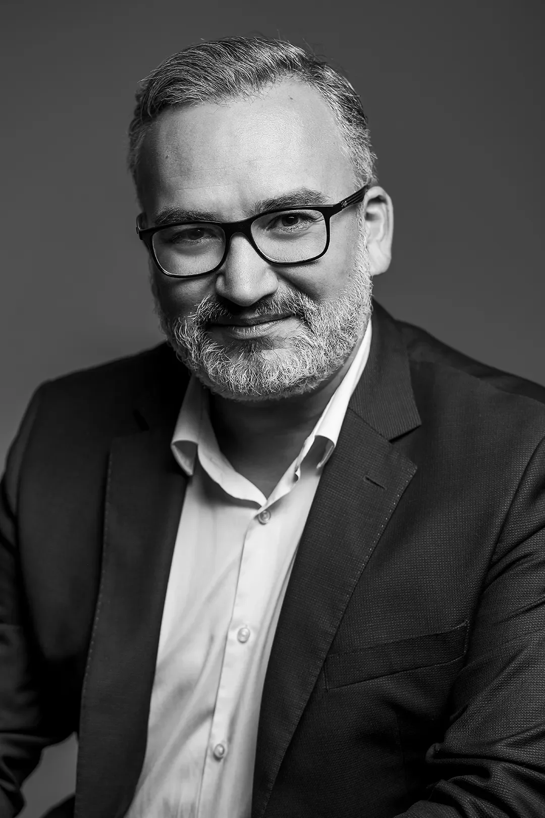 Photo en noir et blanc d'un homme portant des lunettes, ayant une barbe grise, habillé d'une chemise blanche et d'une veste sombre, posé de face avec un léger sourire.