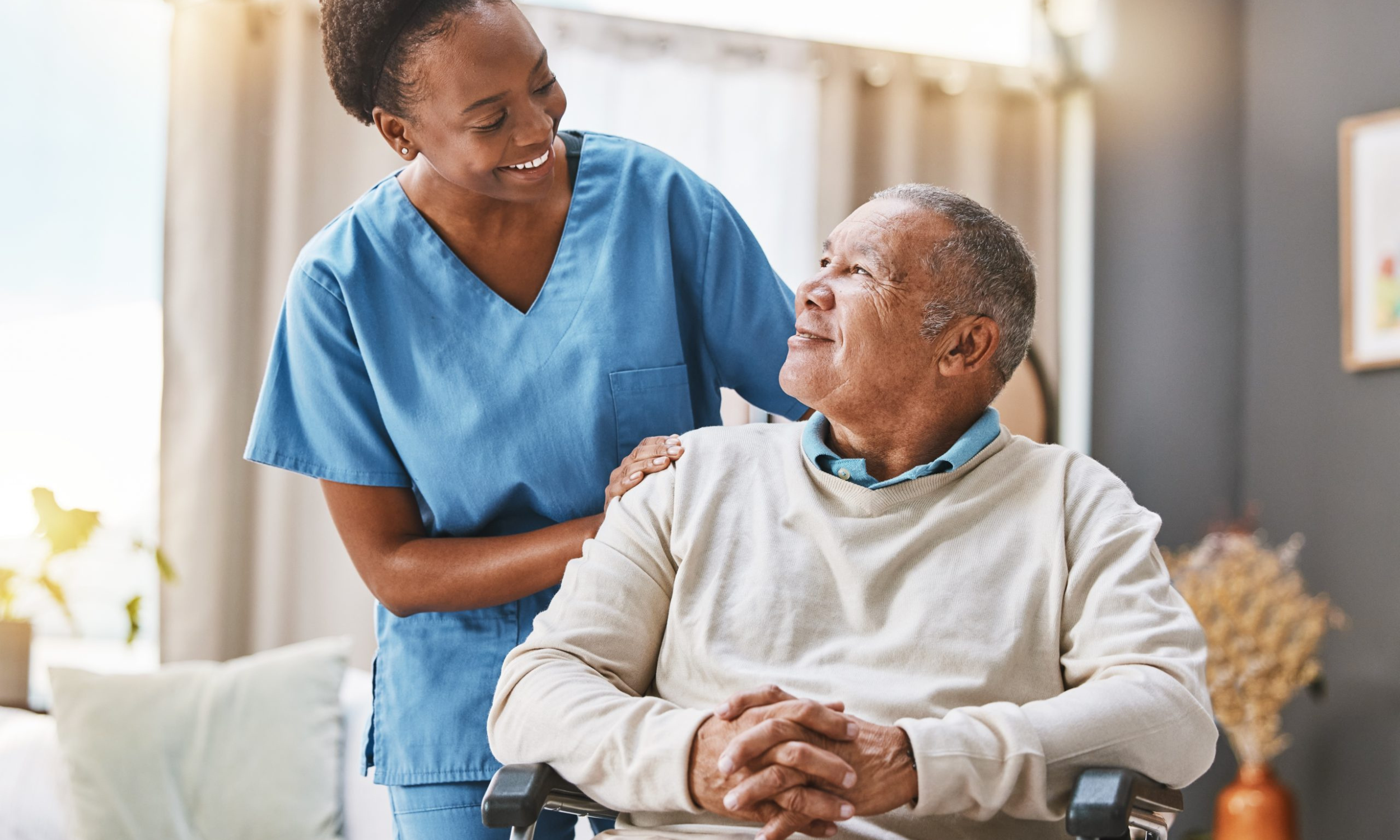 A female healthcare worker in blue scrubs smiling warmly at an elderly man seated in a wheelchair, holding his arm in a caring gesture in a bright, cozy room.