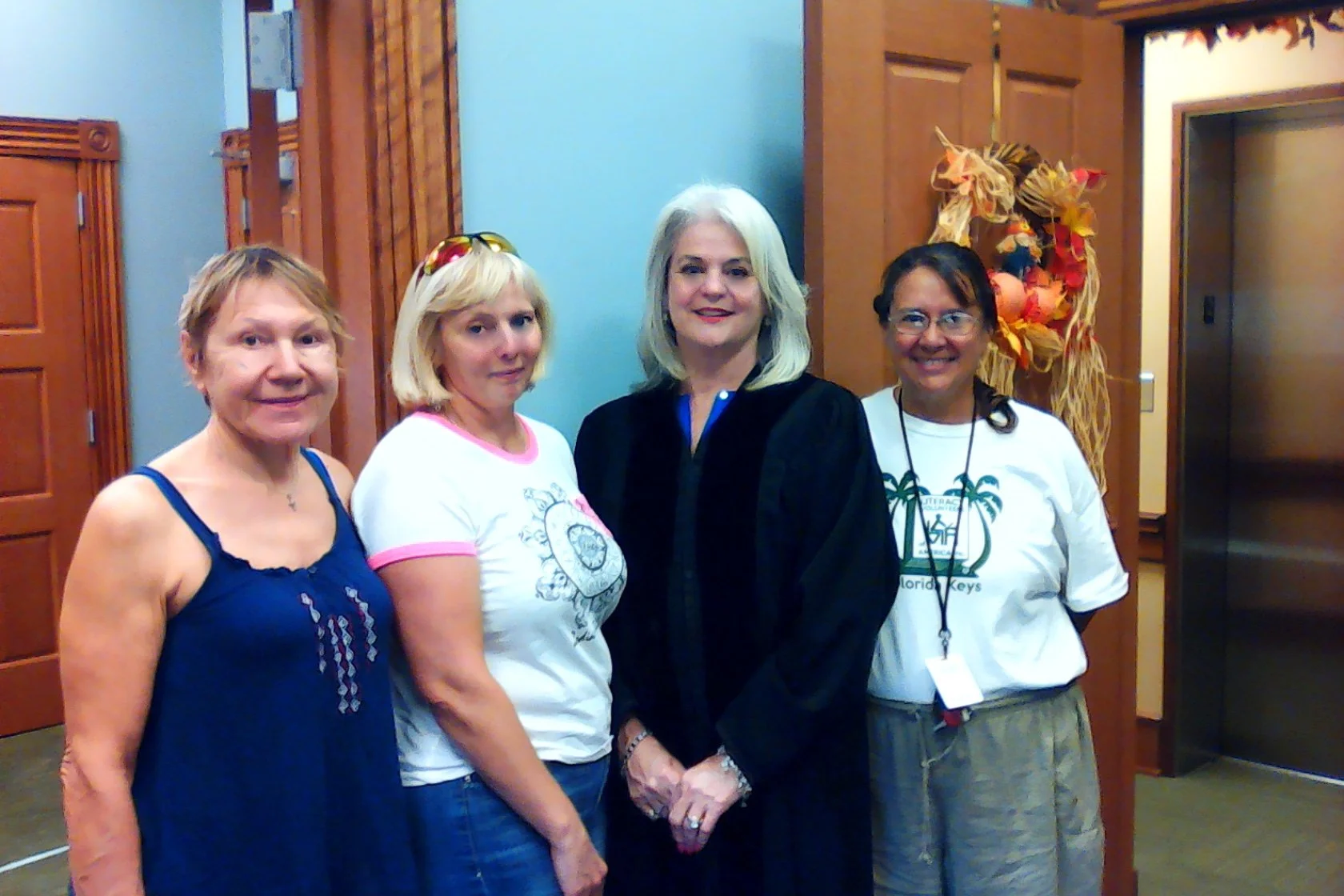 Four women standing together indoors, two with glasses, one in a graduation robe, smiling.