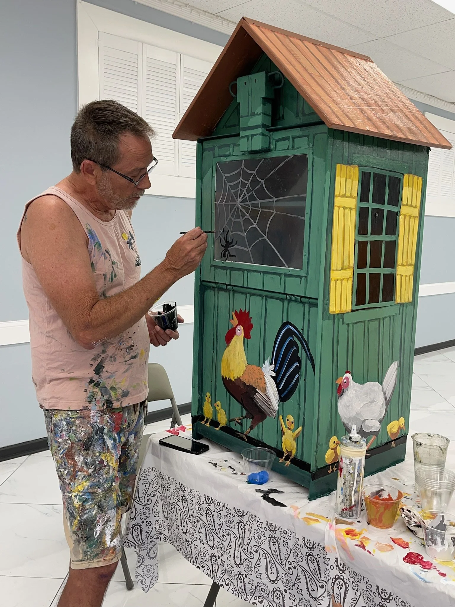 An artist painting a large wooden chicken coop decorated with farm animal artwork, including chickens and ducklings, in an indoor setting.