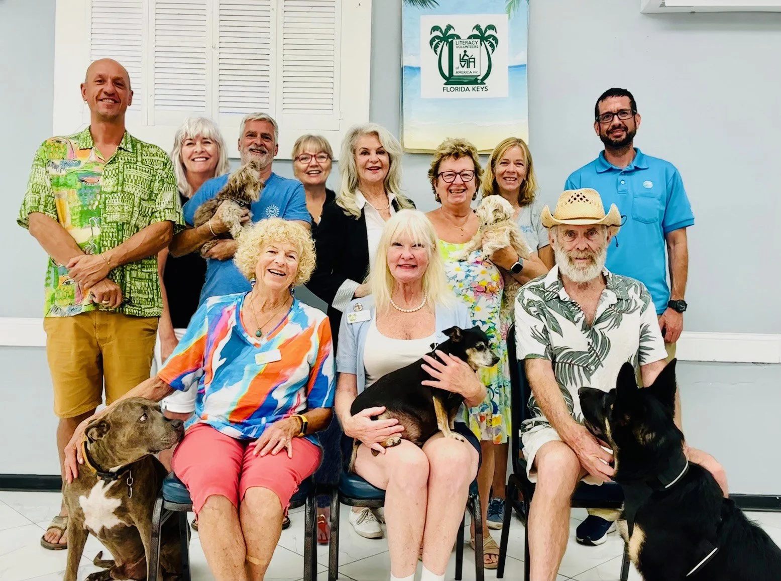 A group of people, including elderly and two men with dogs, gathered in a room with a sign that reads 'Literacy Volunteers Florida Keys.' They are smiling and wearing colorful casual clothing.