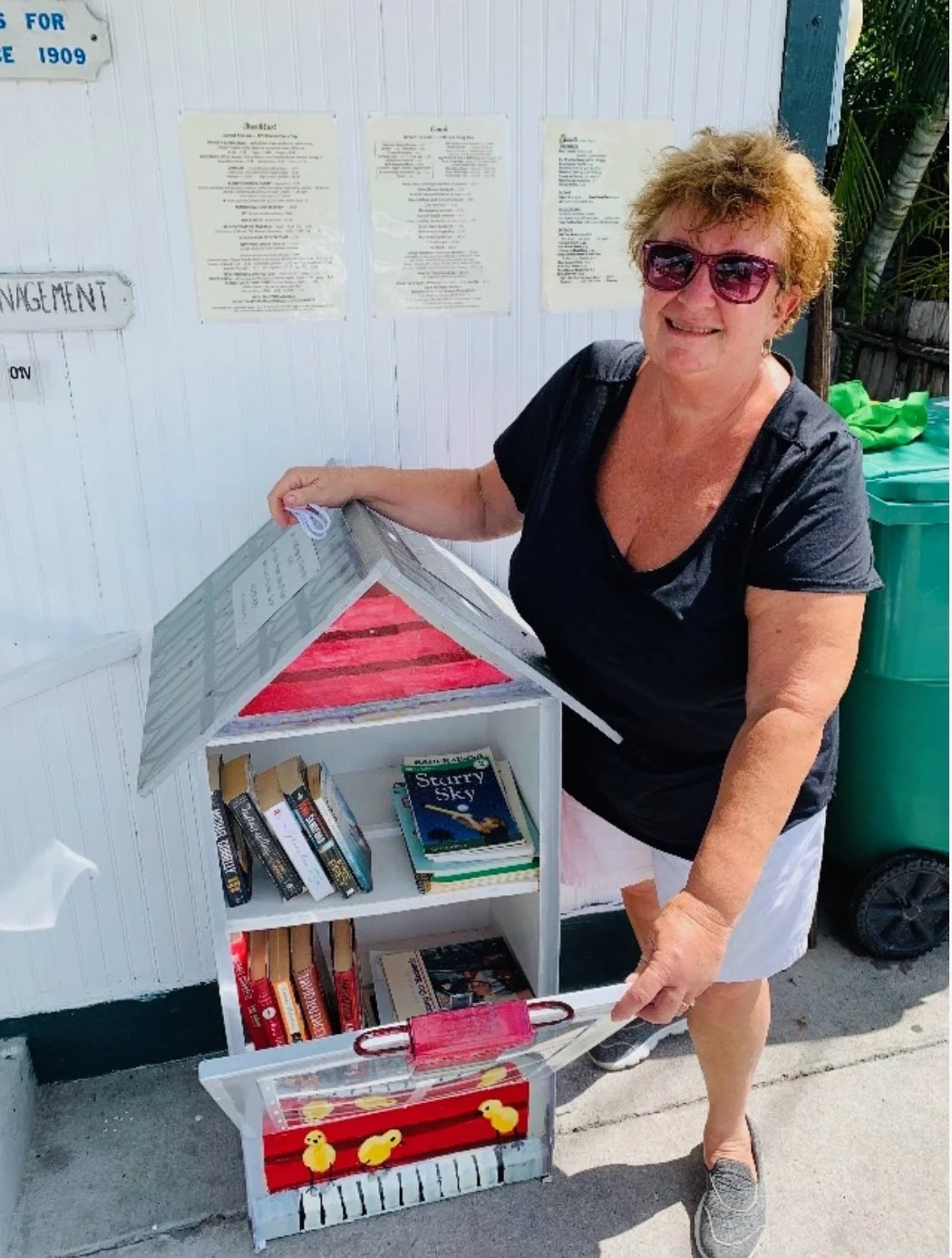 A smiling woman with curly hair, sunglasses, and a black shirt, is opening a small white bookshelf with a pink roof, filled with books, outdoors near a white wall and green garbage bin.