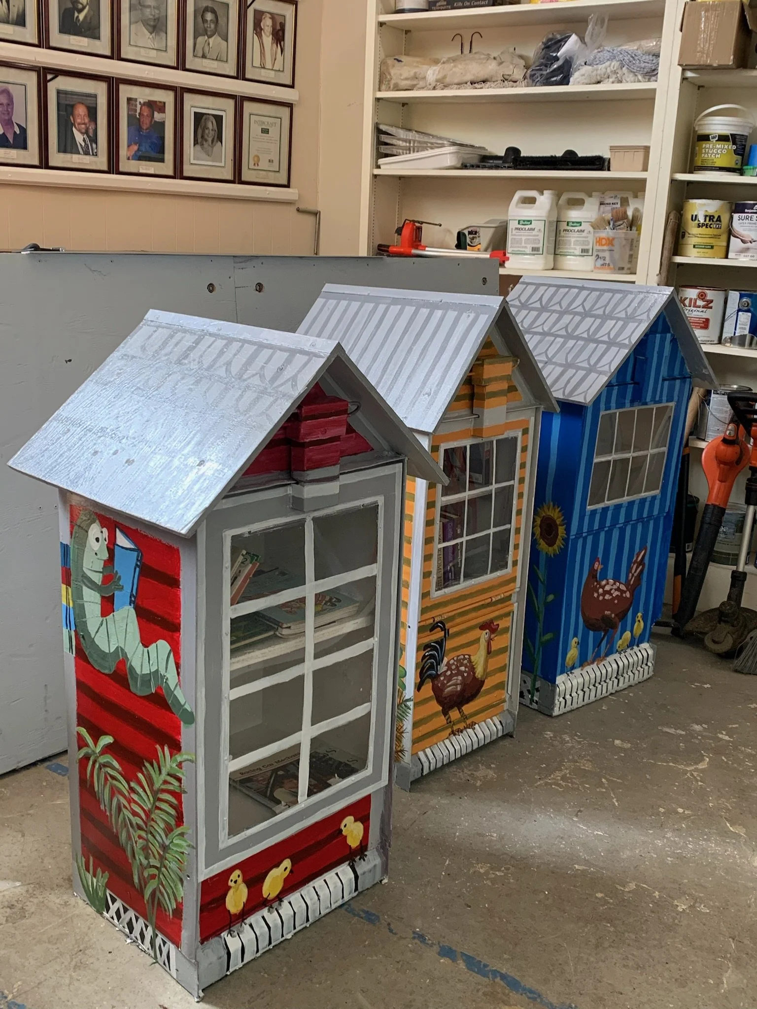 Three colorful painted birdhouses with transparent front panels, decorated with farm animal and nature motifs, situated in a workshop environment.