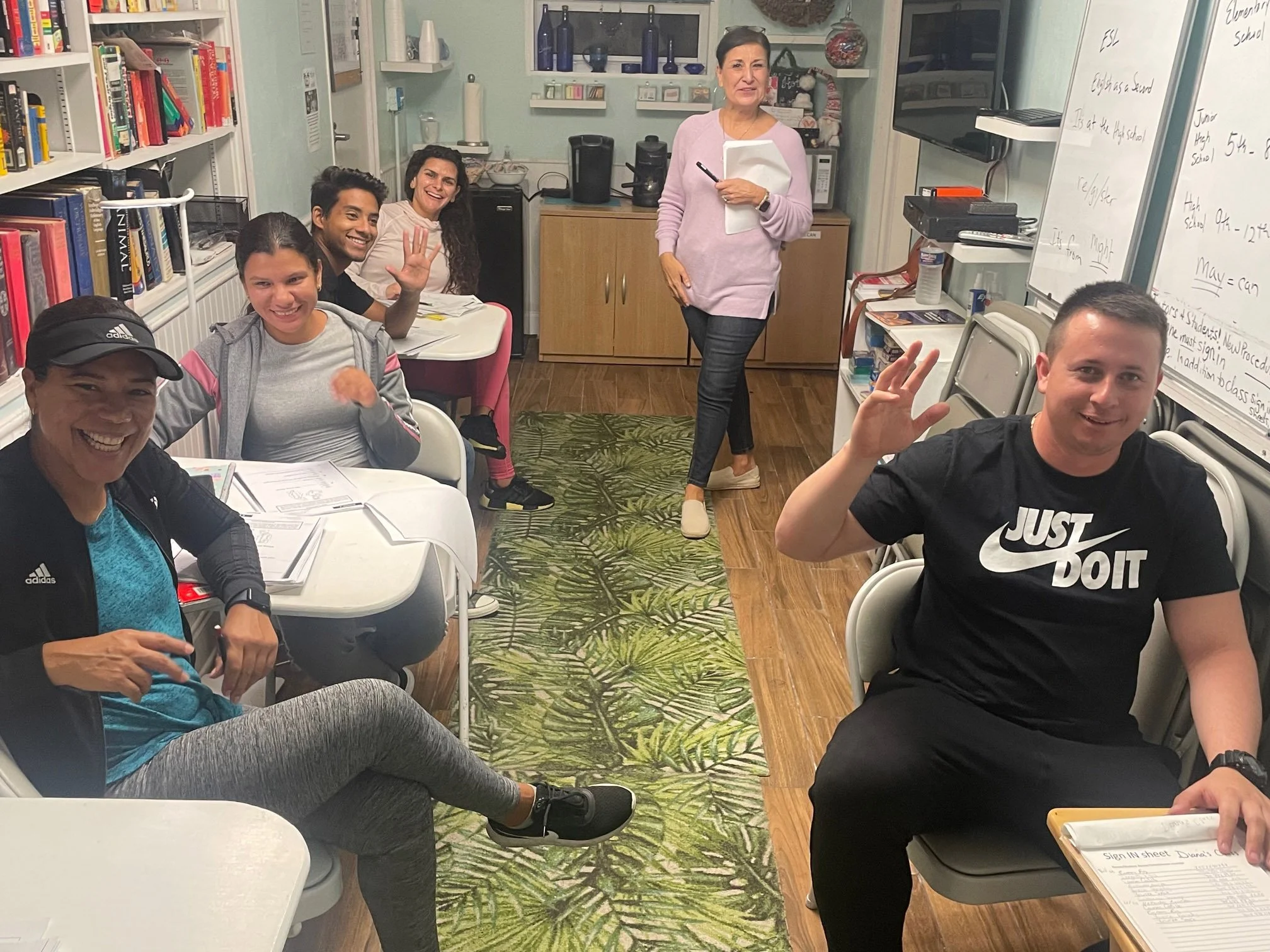 A diverse group of six students sitting in a classroom with a woman teacher standing in the back near a whiteboard. The students are smiling, some are waving, one is holding a clipboard, and others have notebooks. The room has bookshelves, an area with kitchen appliances, and a patterned green rug on the wooden floor.