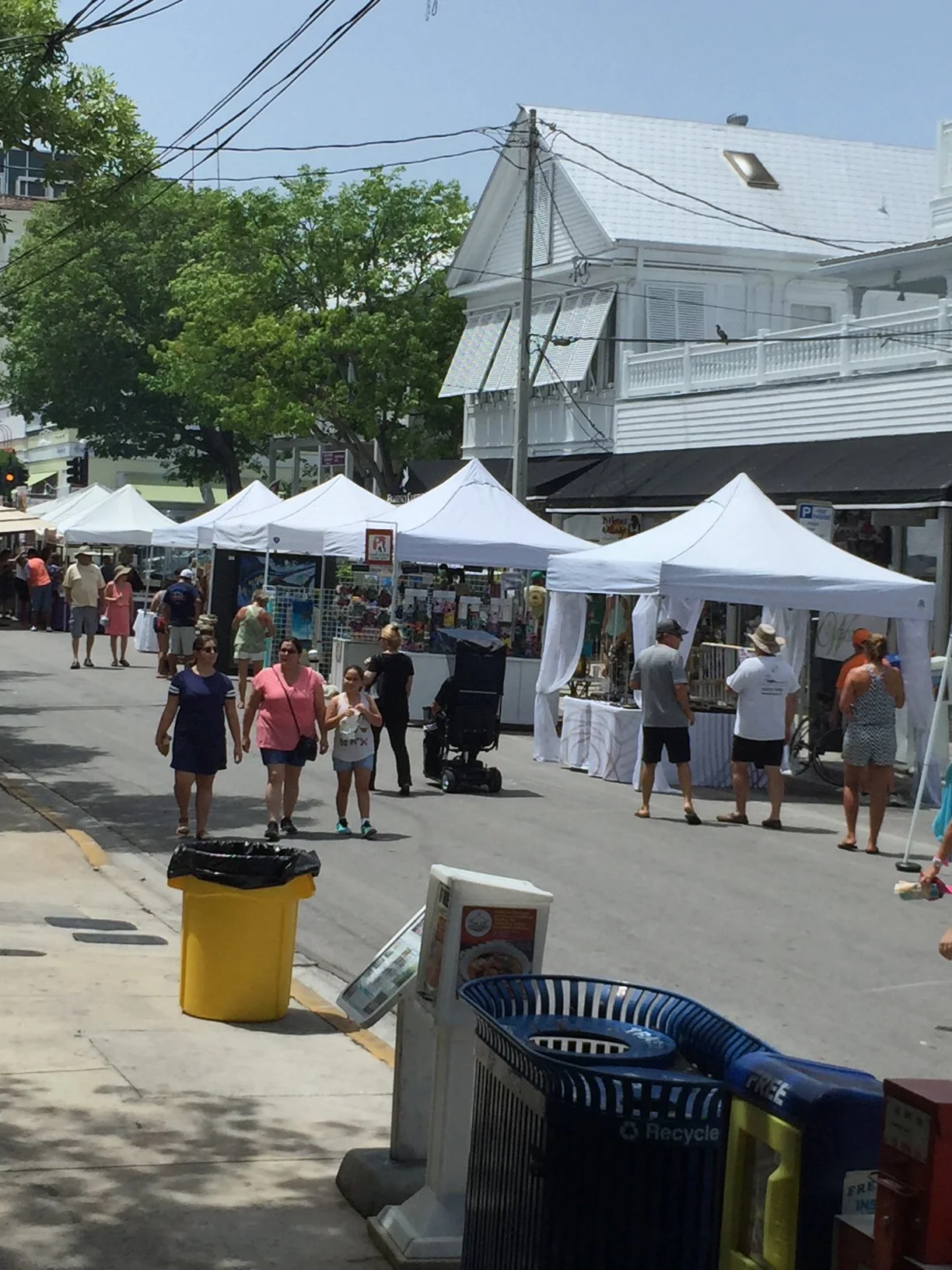 Outdoor street market with white tents and shoppers browsing on a sunny day, with a white building and green trees in the background.