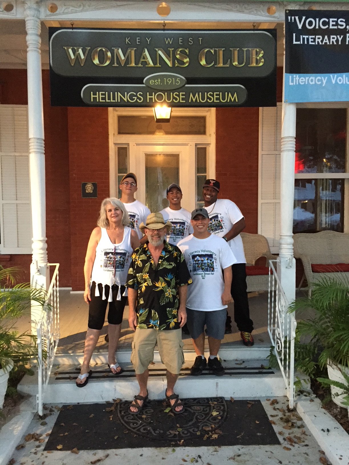 Group of six people standing on the front porch of a historic house in front of a sign that reads 'Key West Women's Club, est. 1915, Helligs House Museum.' The group includes men and women of different ages, some wearing white T-shirts with a logo and text, and others in casual or tropical clothing.