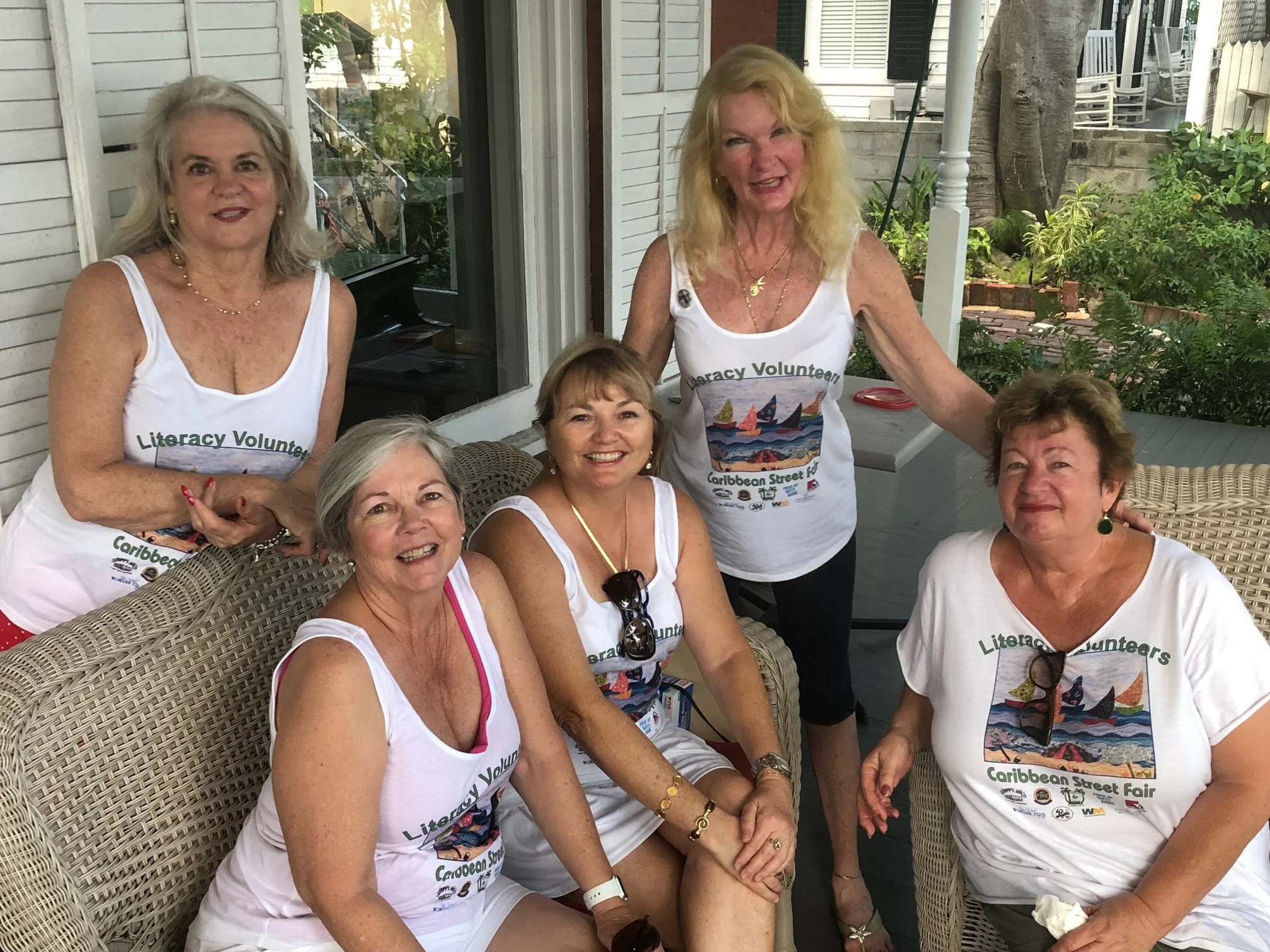 Five women wearing white t-shirts with colorful sailboat and wave graphic, seated and standing on a porch, smiling at the camera, with greenery and outdoor furniture in the background.