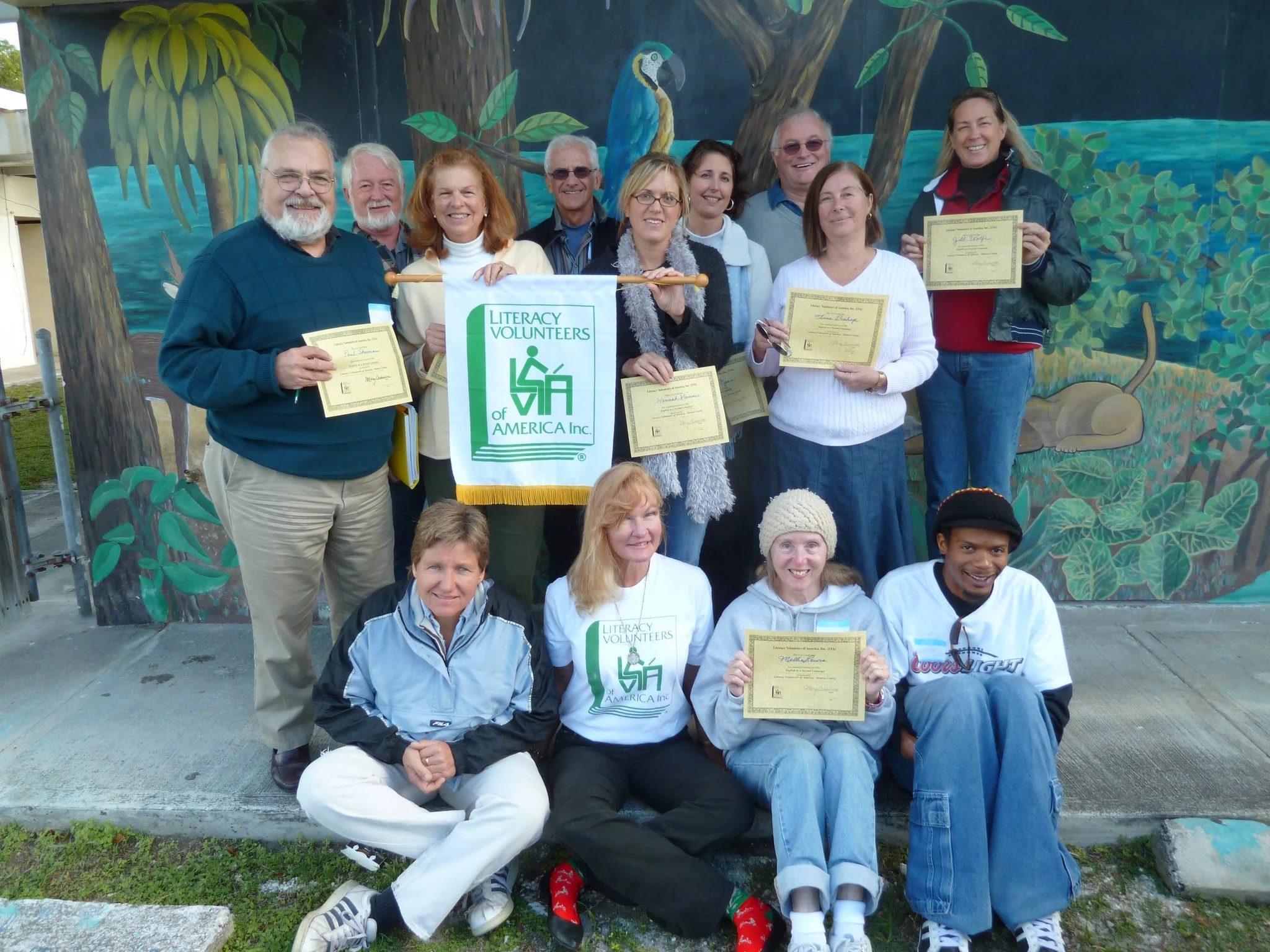 Group of people holding certificates and a Literacy Volunteers of America banner, standing in front of a colorful mural with trees and a parrot painted on the wall.