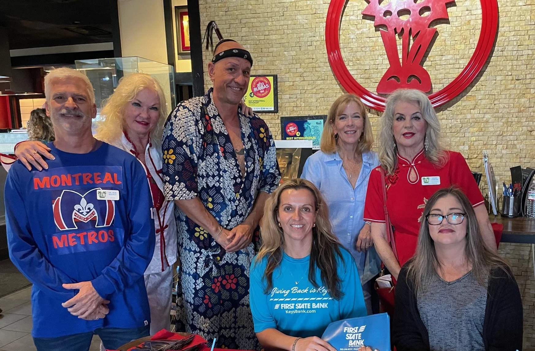 Group of seven people posing inside a restaurant. They are smiling, some with their arms around each other. The background includes a brick wall with a large red logo and menus. The group appears to be at a gathering or event.