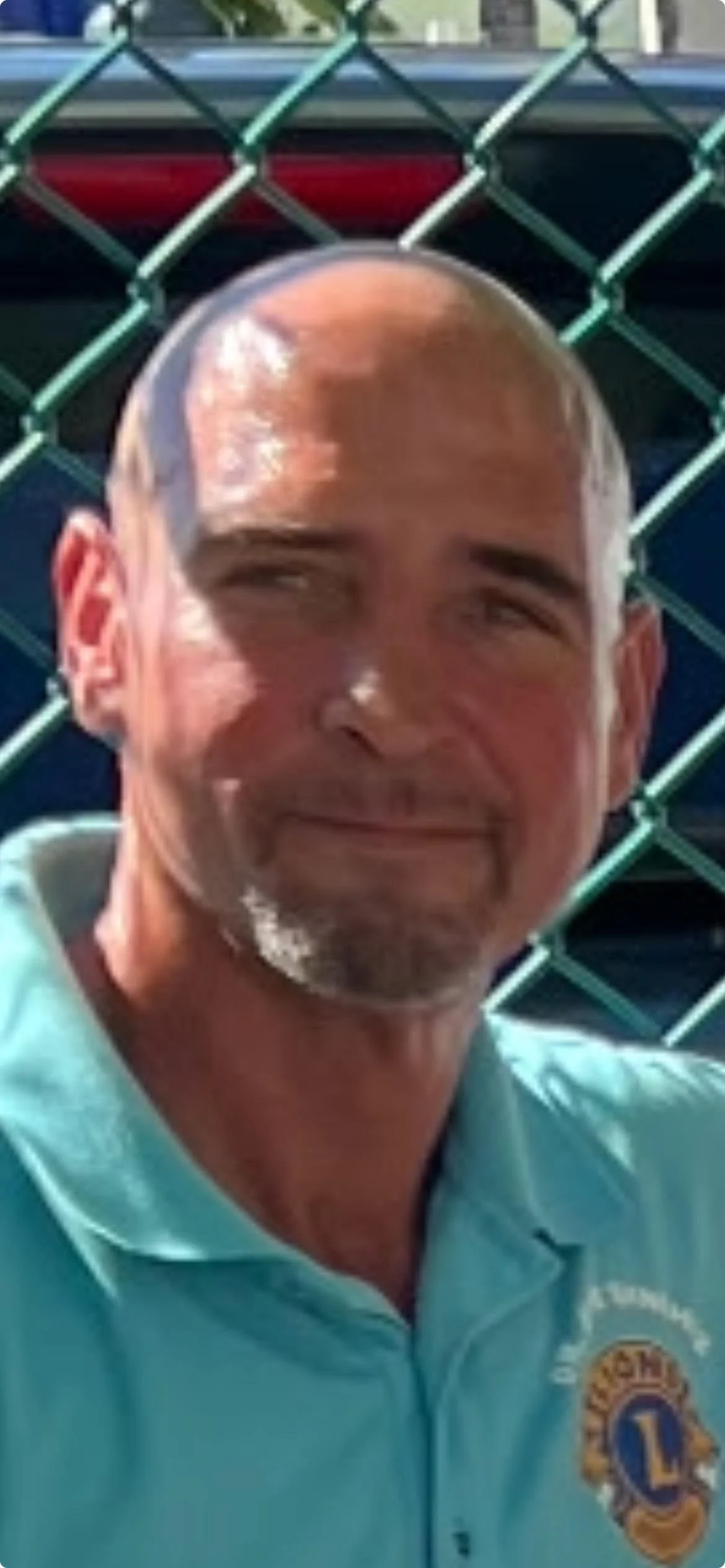 A man with a shaved head and goatee smiling, wearing a light blue shirt with a Lions Club logo, standing outdoors in front of a chain-link fence and a parked vehicle.