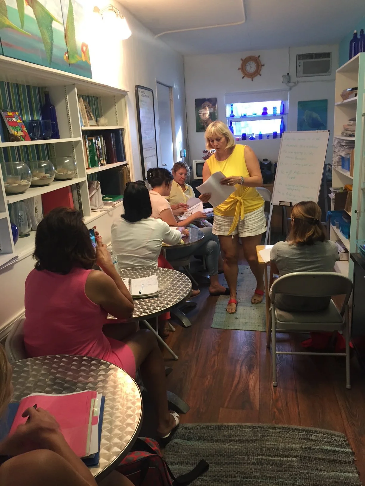 A group of women sitting around small round tables in a cozy room, with a woman in a yellow top standing near a whiteboard, leading a discussion or presentation.