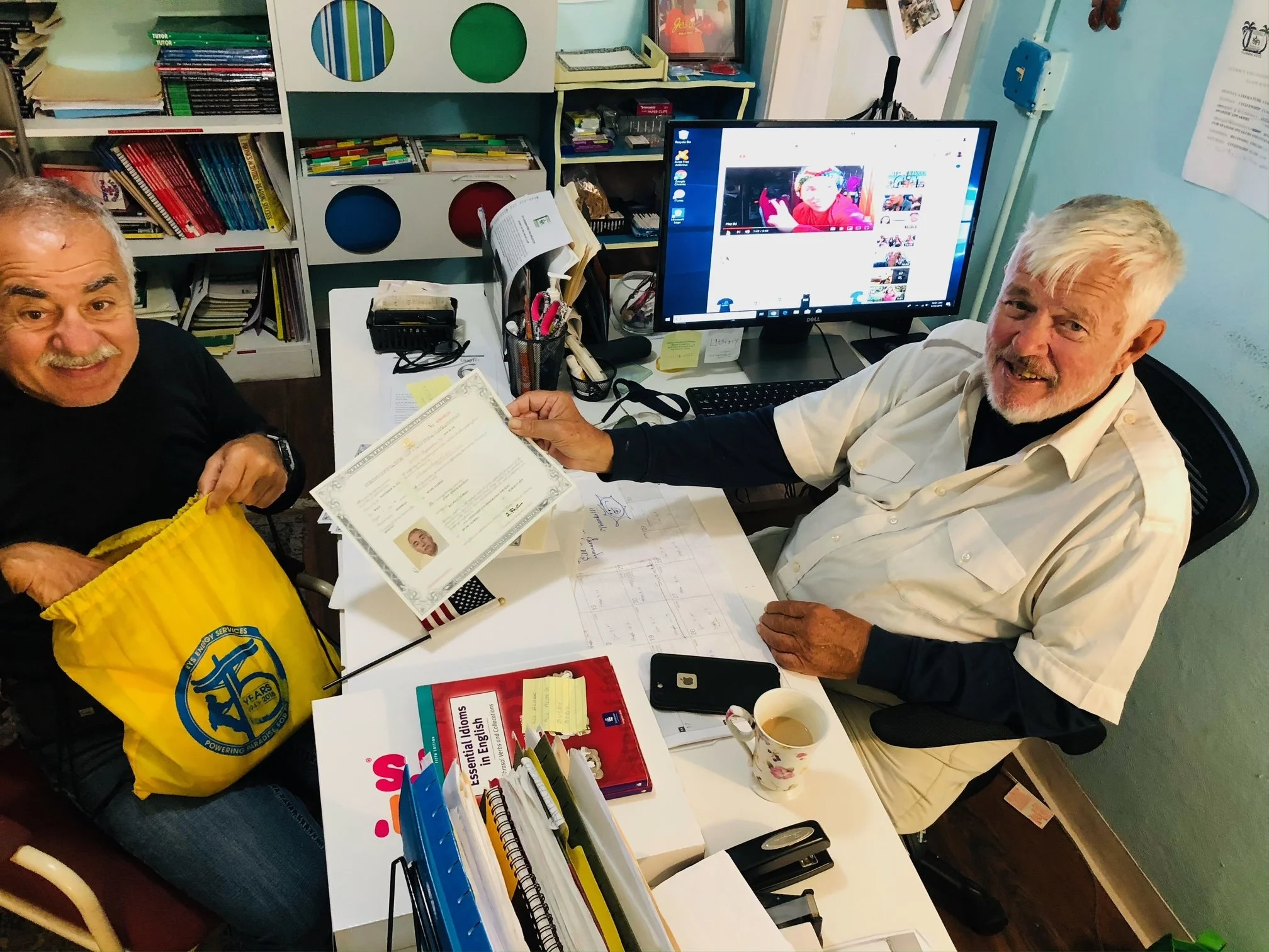 Two elderly men smiling at a desk, one of them holding a certificate. The desk has various office supplies, papers, a computer monitor, and a coffee mug. The background shows bookshelves with colorful books and decorations.
