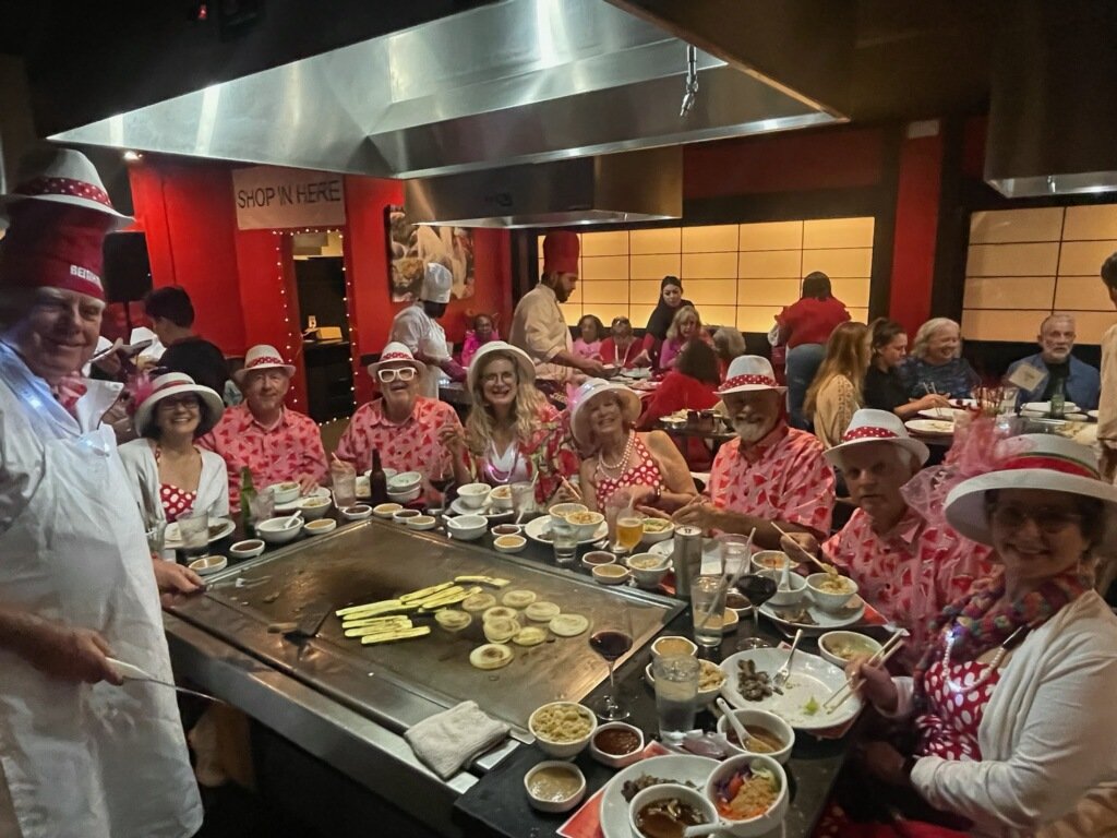 Group of people dressed in red and white, enjoying a teppanyaki dining experience at a restaurant with a large grill at the center, surrounded by bowls of sauces and ingredients.