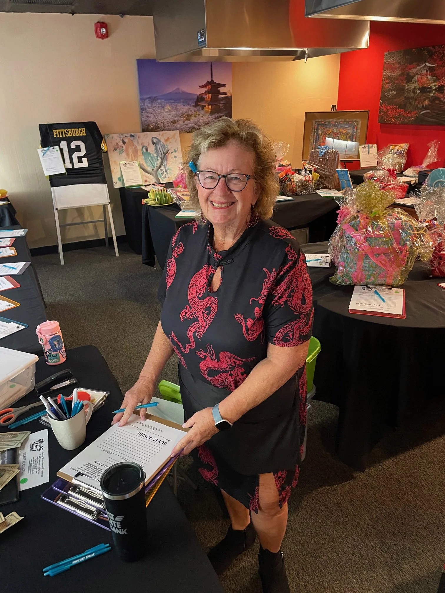 A woman standing at a table filled with papers and office supplies, smiling at the camera. The background features various artwork, gift baskets, and a Pittsburgh Steelers jersey with the number 12.