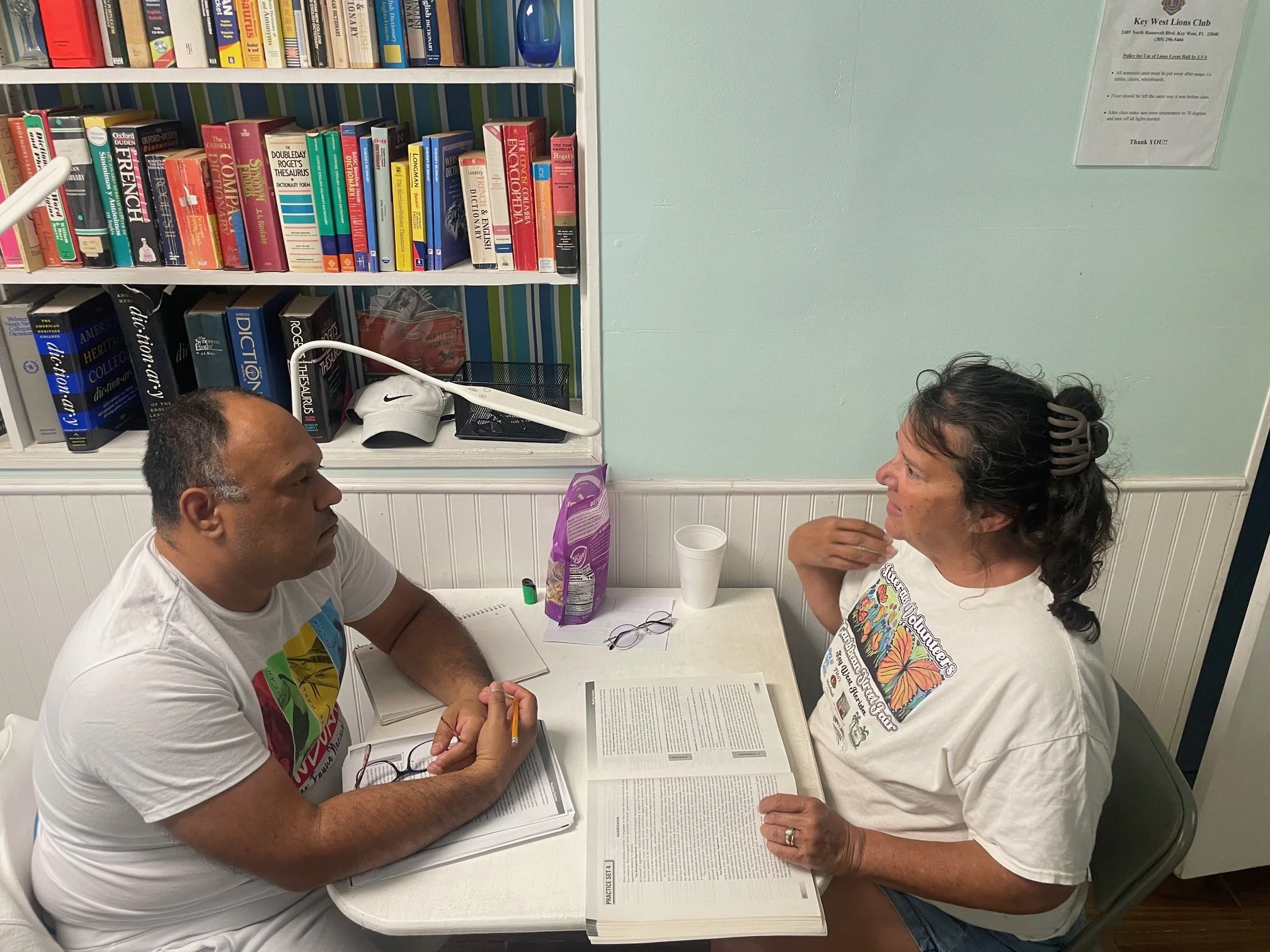 A man and woman are sitting at a table, engaging in a serious conversation. The man has short hair and is wearing a white T-shirt with colorful graphics. The woman has curly hair held back with a hair clip and is wearing a white T-shirt with colorful butterfly print. They have open books and papers in front of them.