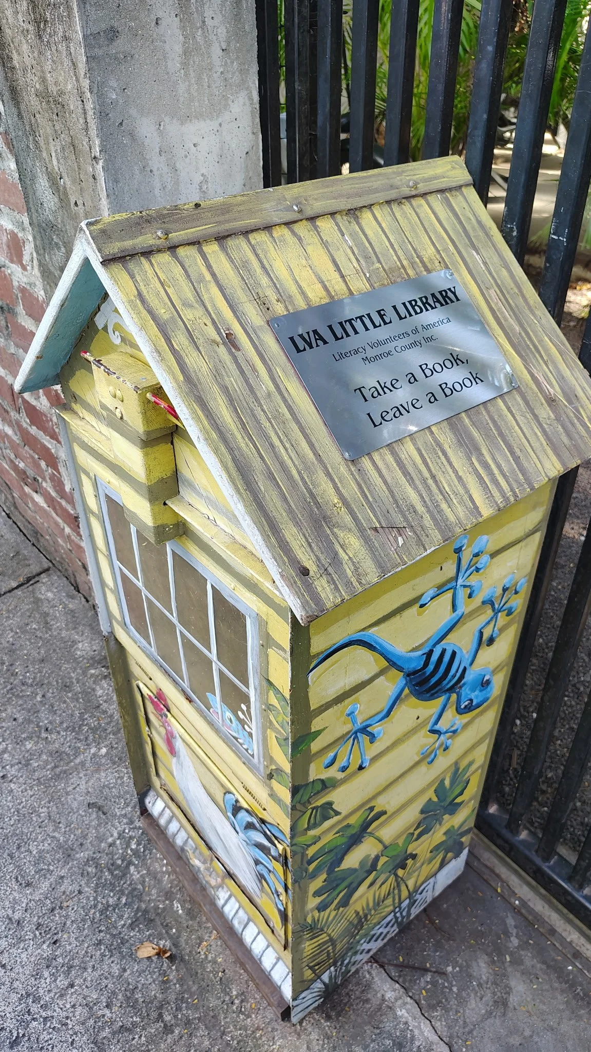 Decorative community library box painted yellow with a blue lizard, white bird, and window designs, mounted on a sidewalk near brick and metal fencing.