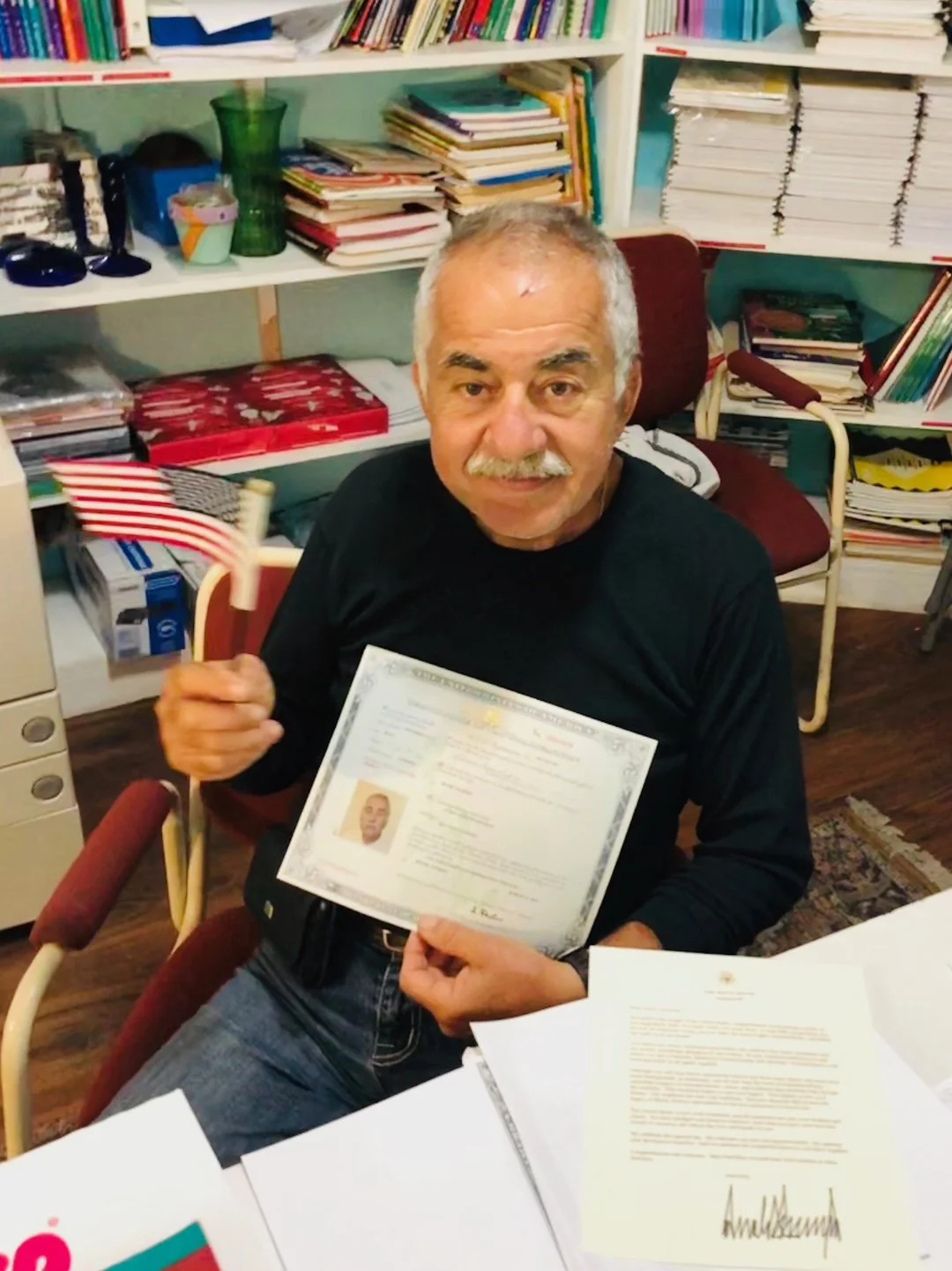 An older man sitting at a desk holding a certificate in one hand and a small American flag in the other. The background shows shelves filled with books and papers, indicating an office or study environment.