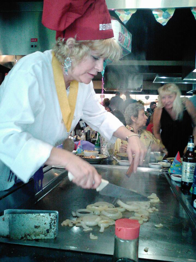A woman wearing a red chef hat and white coat is cooking onions on a griddle at a party, with people and party decorations in the background.