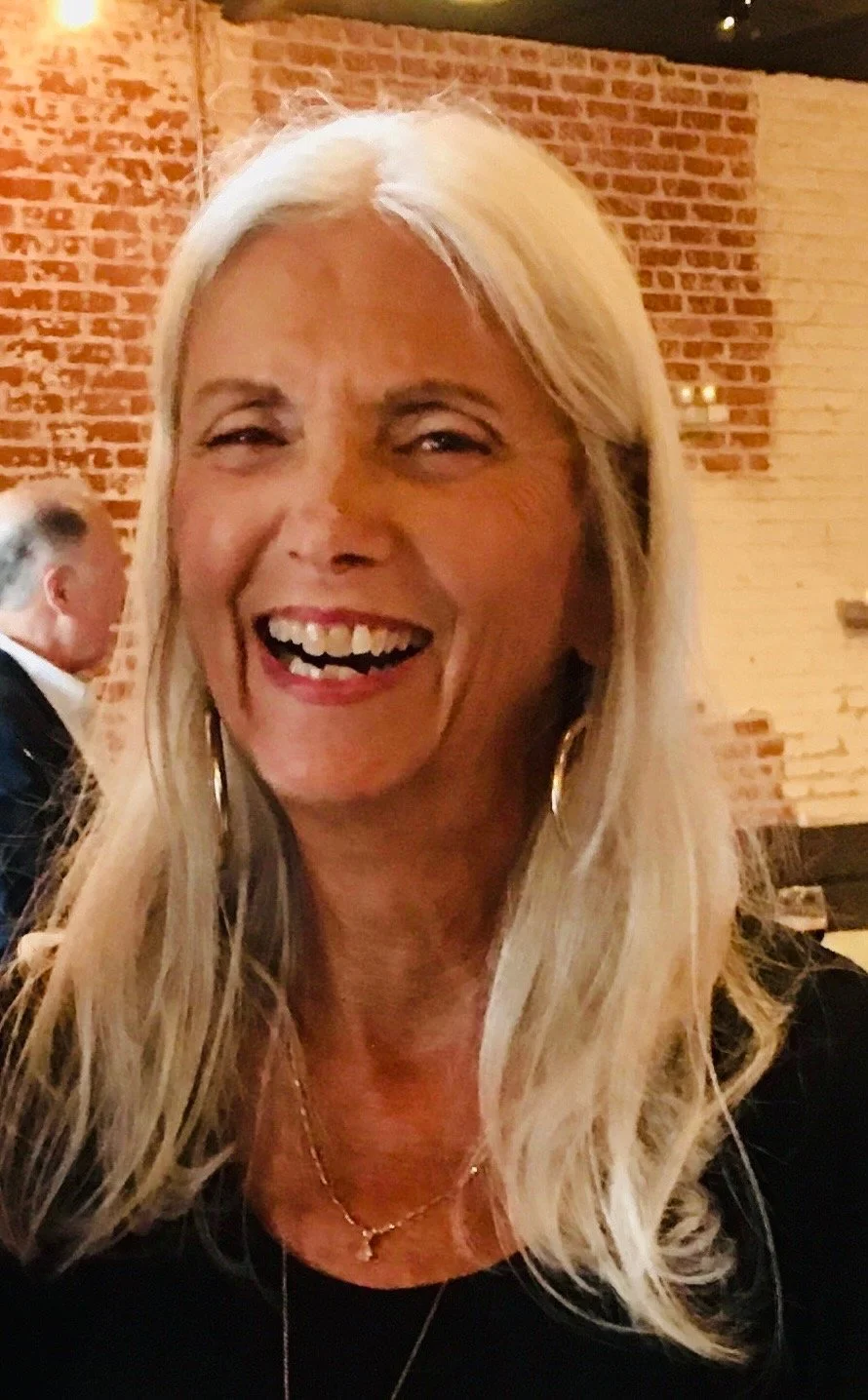 A smiling woman with long blonde hair, wearing earrings and a necklace, in a restaurant with brick walls.