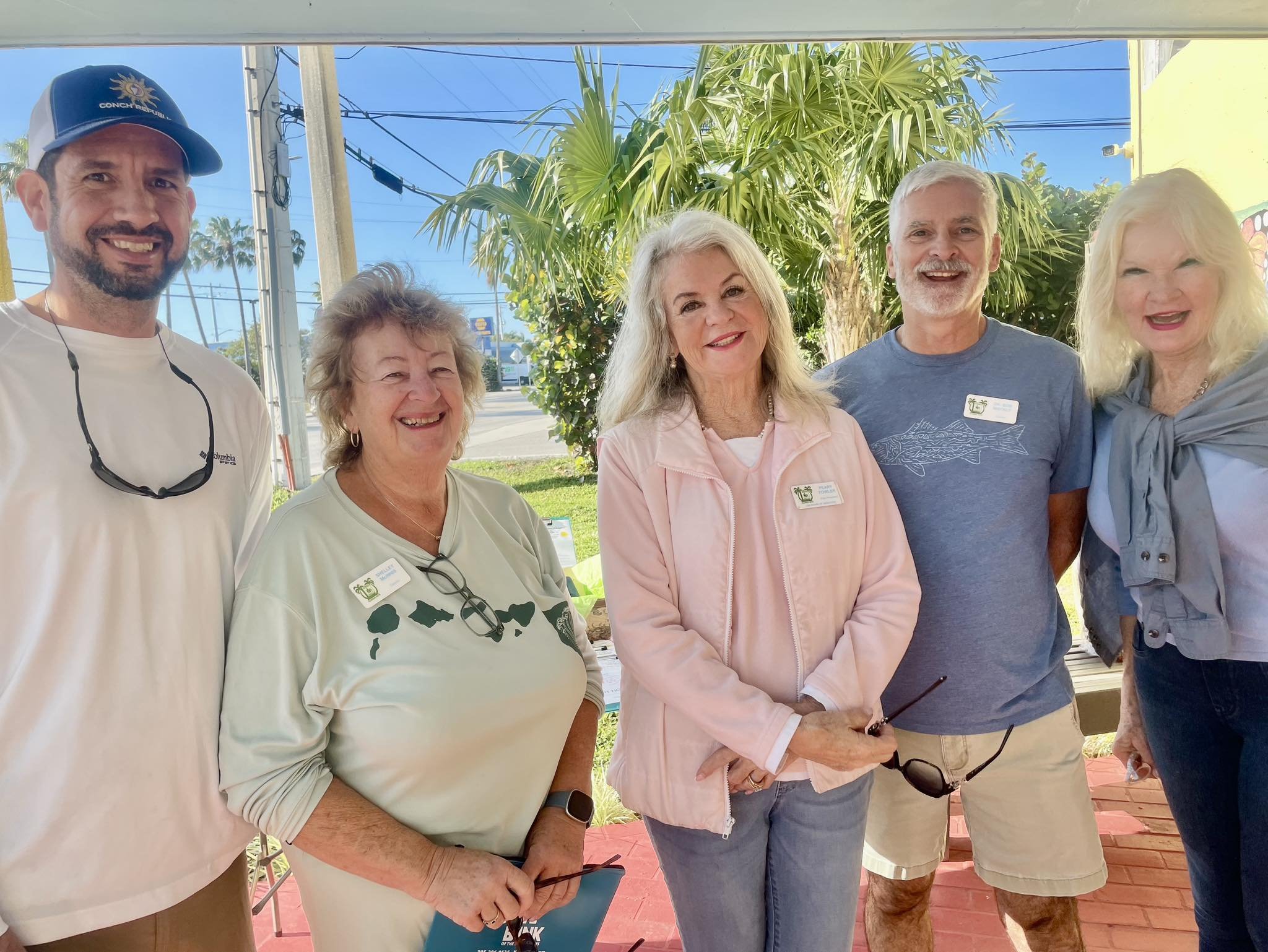 Five smiling people standing outdoors in front of greenery, with a bright blue sky and power lines behind them. They are wearing casual clothes and name tags, suggesting a social or community gathering.