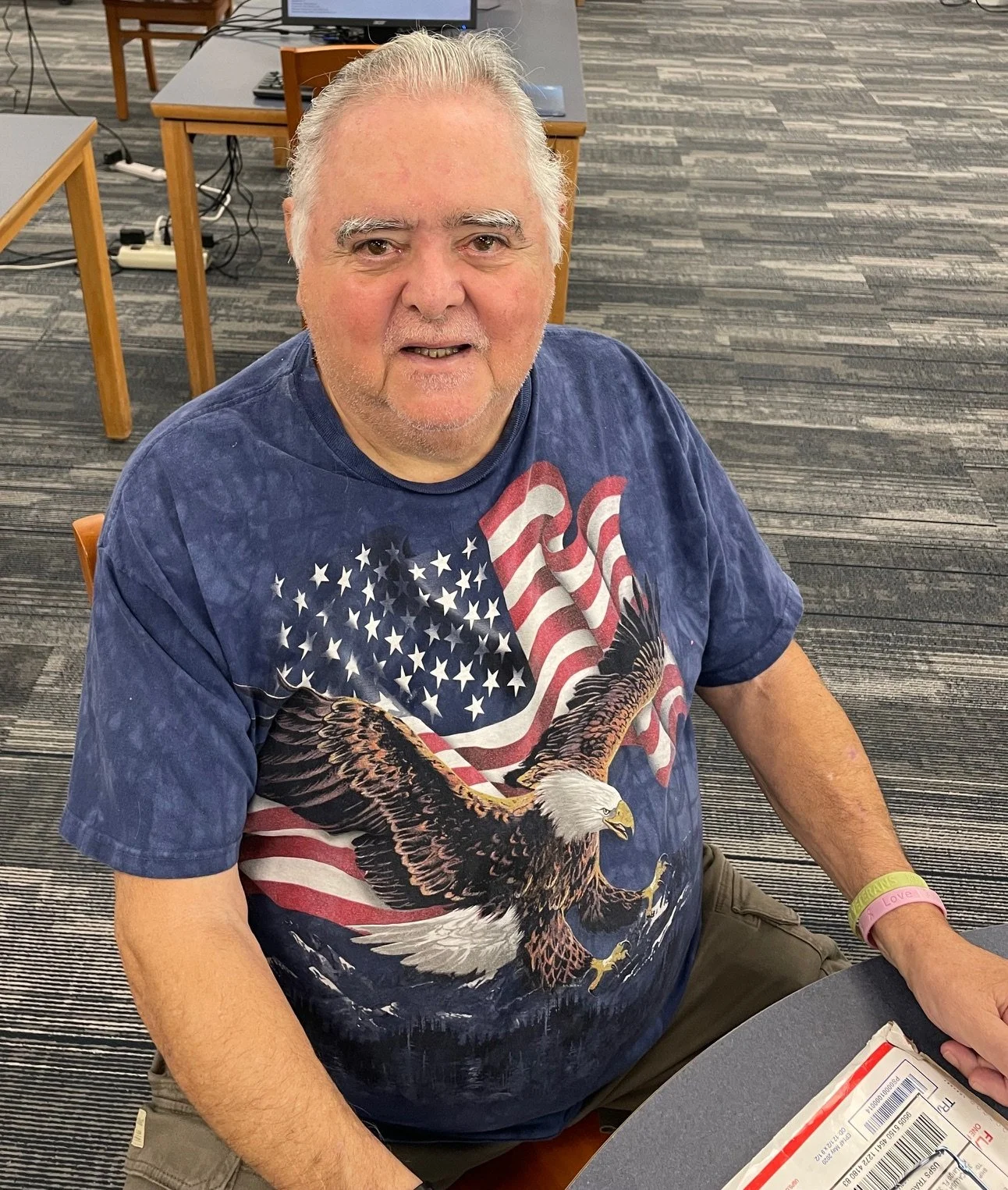 An older man with gray hair and a beard sitting at a table, wearing a navy blue t-shirt with a large design of an American flag and a bald eagle.