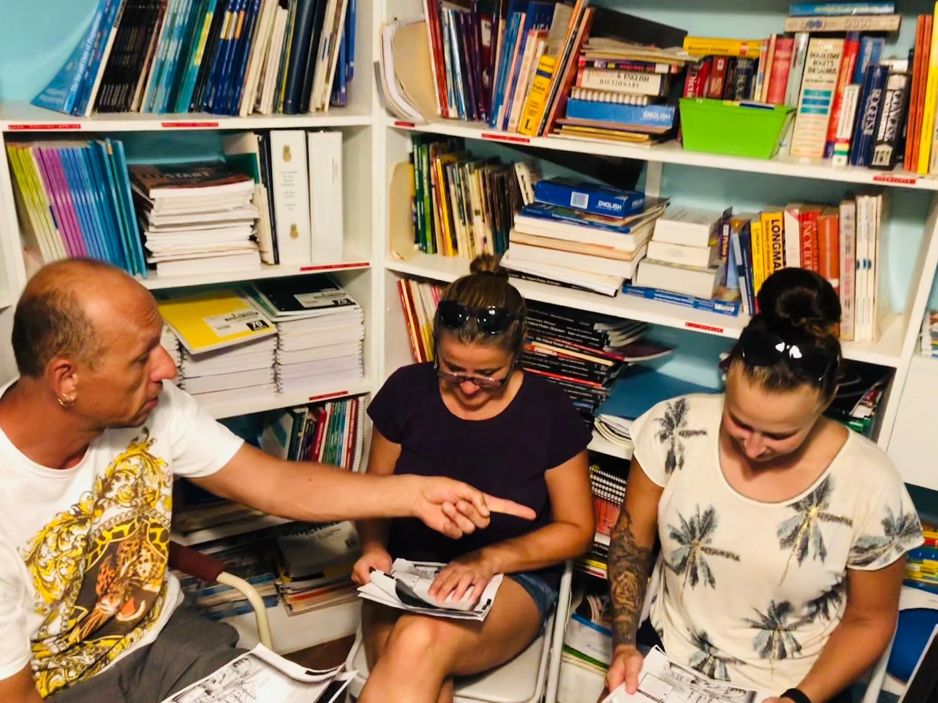 Three people sitting in a room filled with books, engaged in a discussion, with one person pointing at a book or paper.