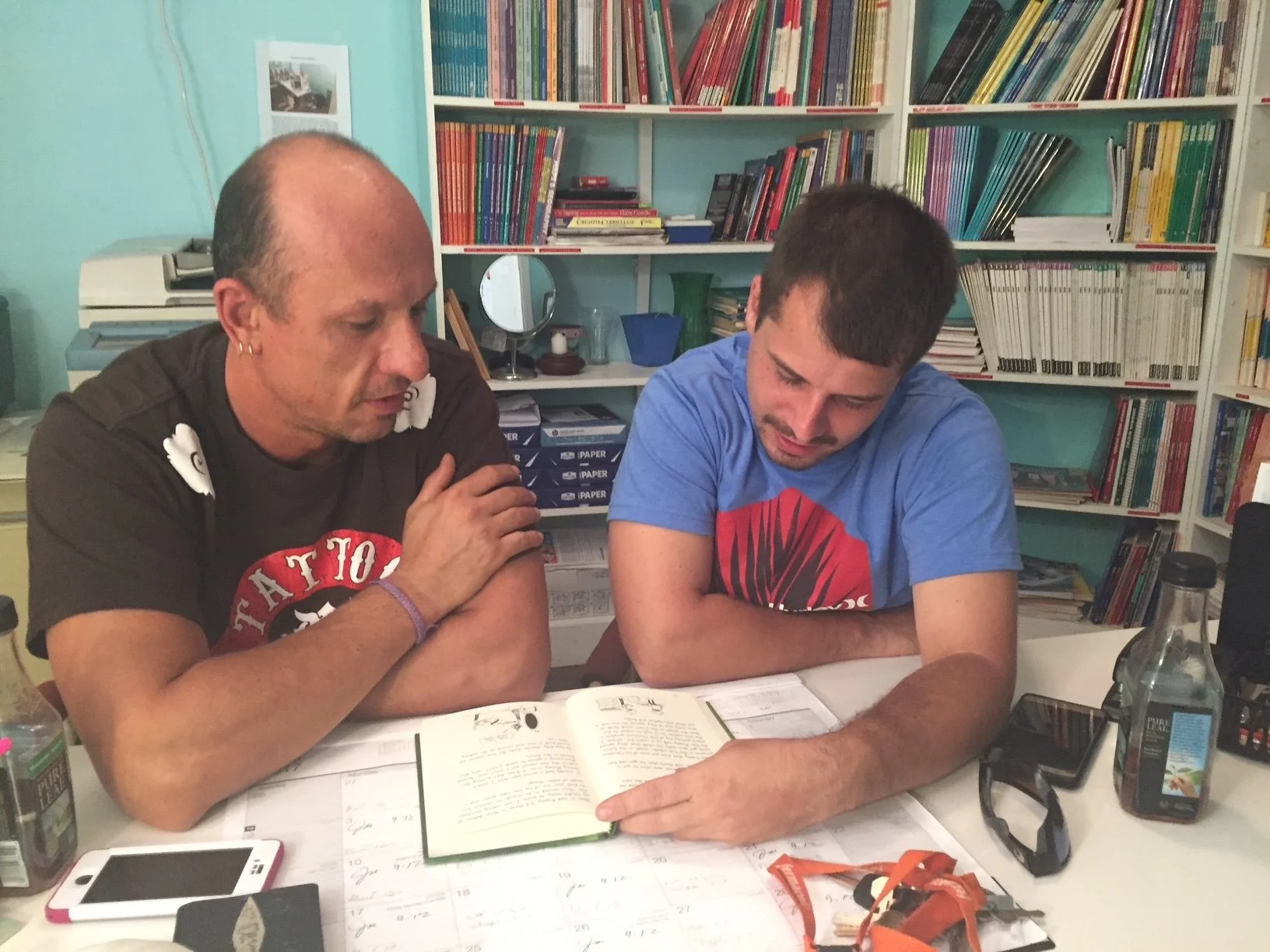 Two men sitting at a table, reading and discussing a book, with shelves filled with books and office supplies behind them.
