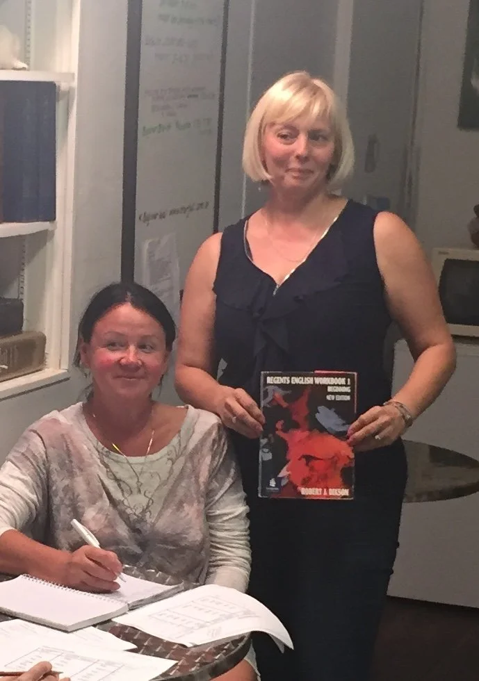 Two women in an office or classroom setting, one sitting at a table with notebooks and papers, the other standing and holding a book titled 'Records English Workbook 1'.