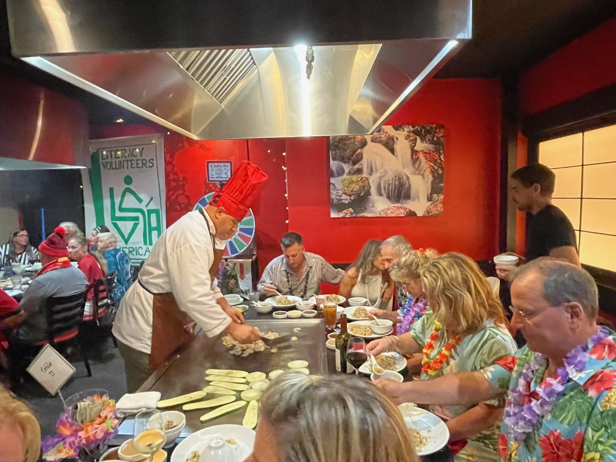 Chef preparing food on a teppanyaki grill in a restaurant with customers seated around, some wearing Hawaiian shirts and leis, and a colorful background with waterfall artwork.