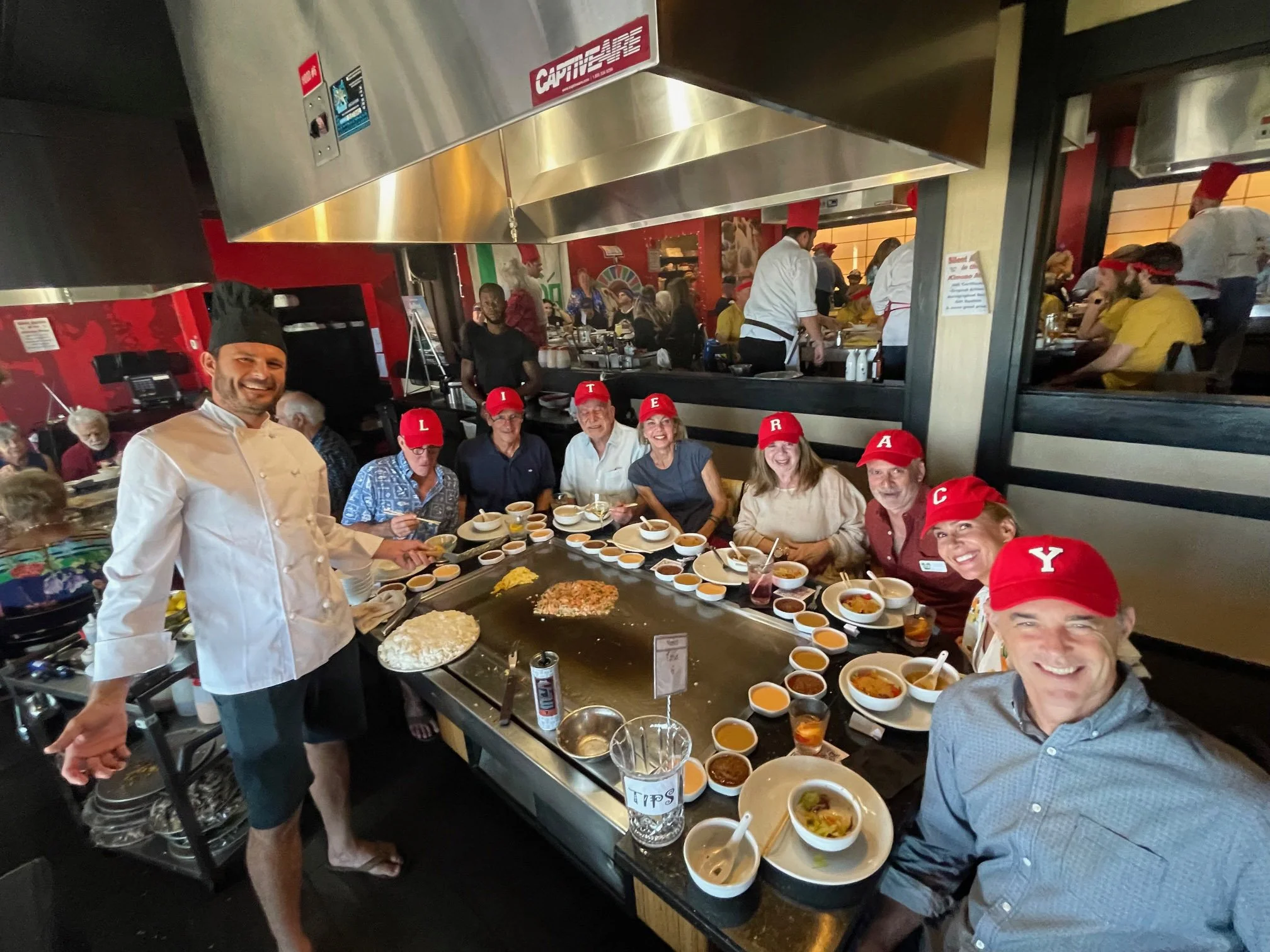 Group of people wearing red caps with letters spelling 'LUNCH' gathered around teppanyaki grill at a restaurant, Chef in white uniform preparing food, other diners in background with vibrant decor.