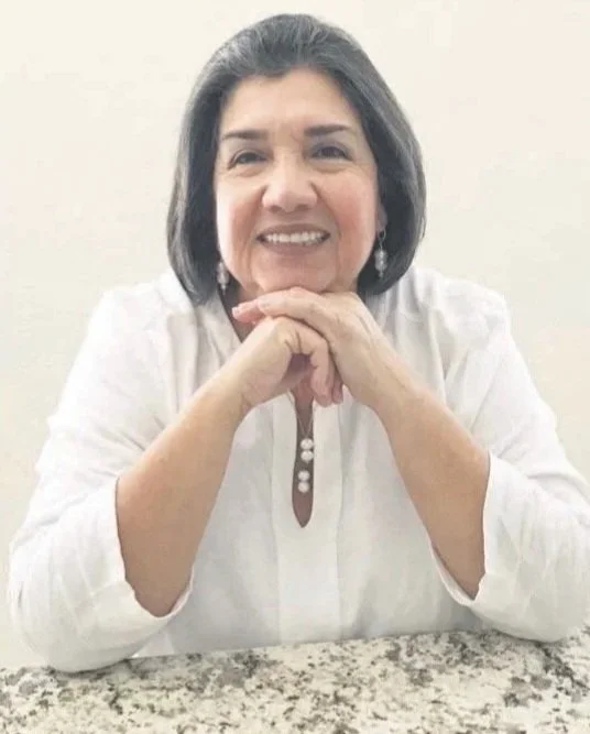A middle-aged woman with shoulder-length dark hair, wearing a white blouse, sitting at a table with her hands clasped under her chin, smiling at the camera against a plain light-colored wall.