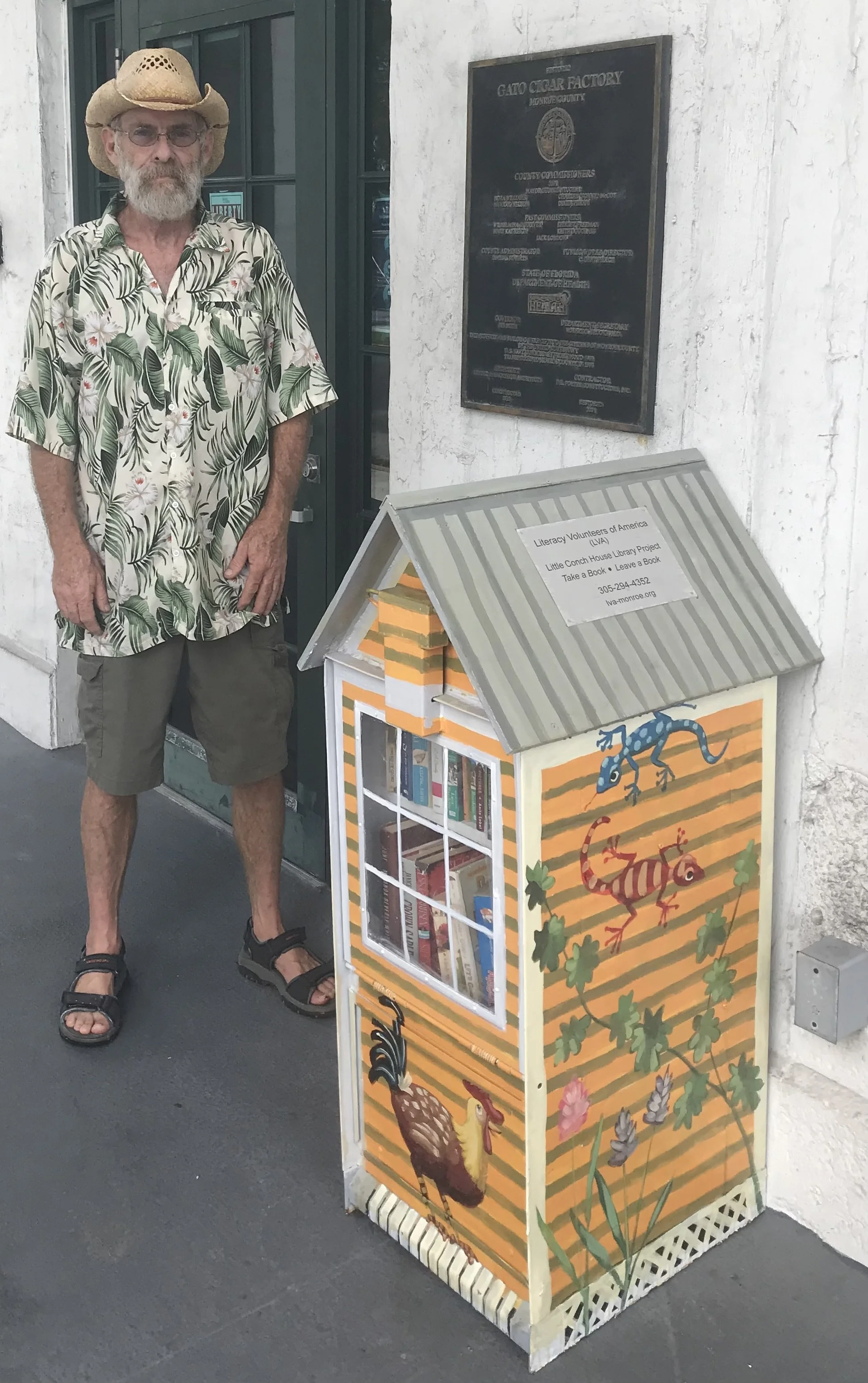 A man standing next to a decorated Little Free Library outside a building. The man is wearing a straw hat, glasses, a tropical shirt, shorts, and sandals. The library is painted with colorful animals and plants.