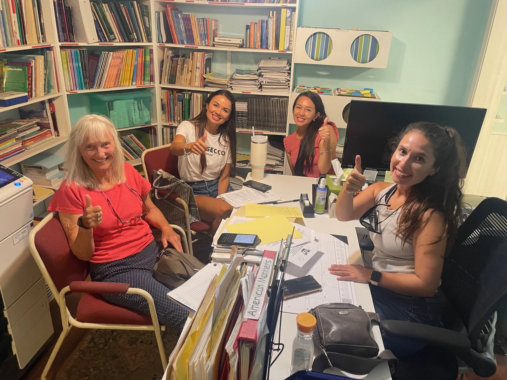 Four women smiling and giving thumbs up around a cluttered desk in an office, with bookshelves filled with books in the background.